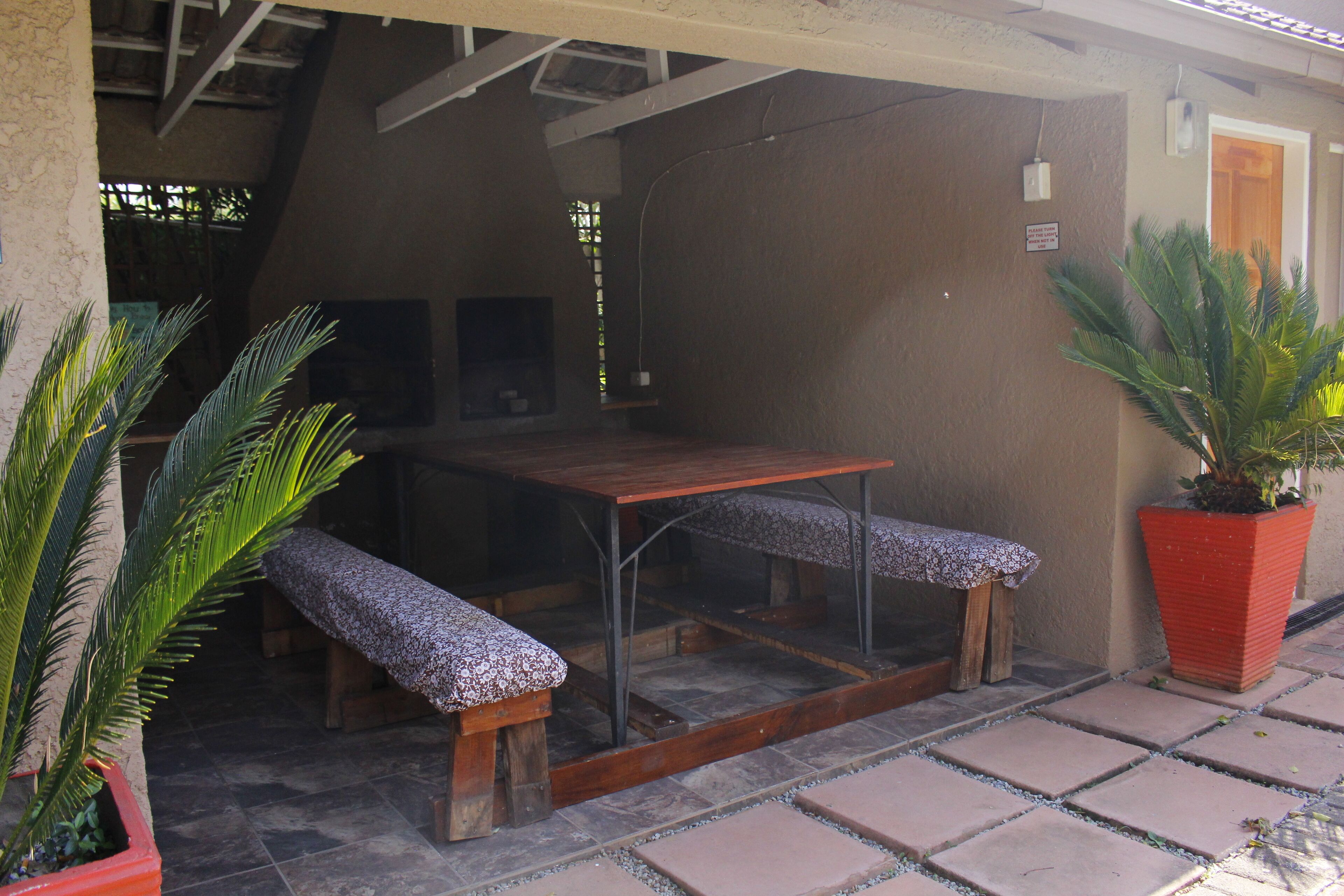 Un patio al aire libre con una mesa de madera y bancos, rodeado de plantas en macetas y un suelo de baldosas.