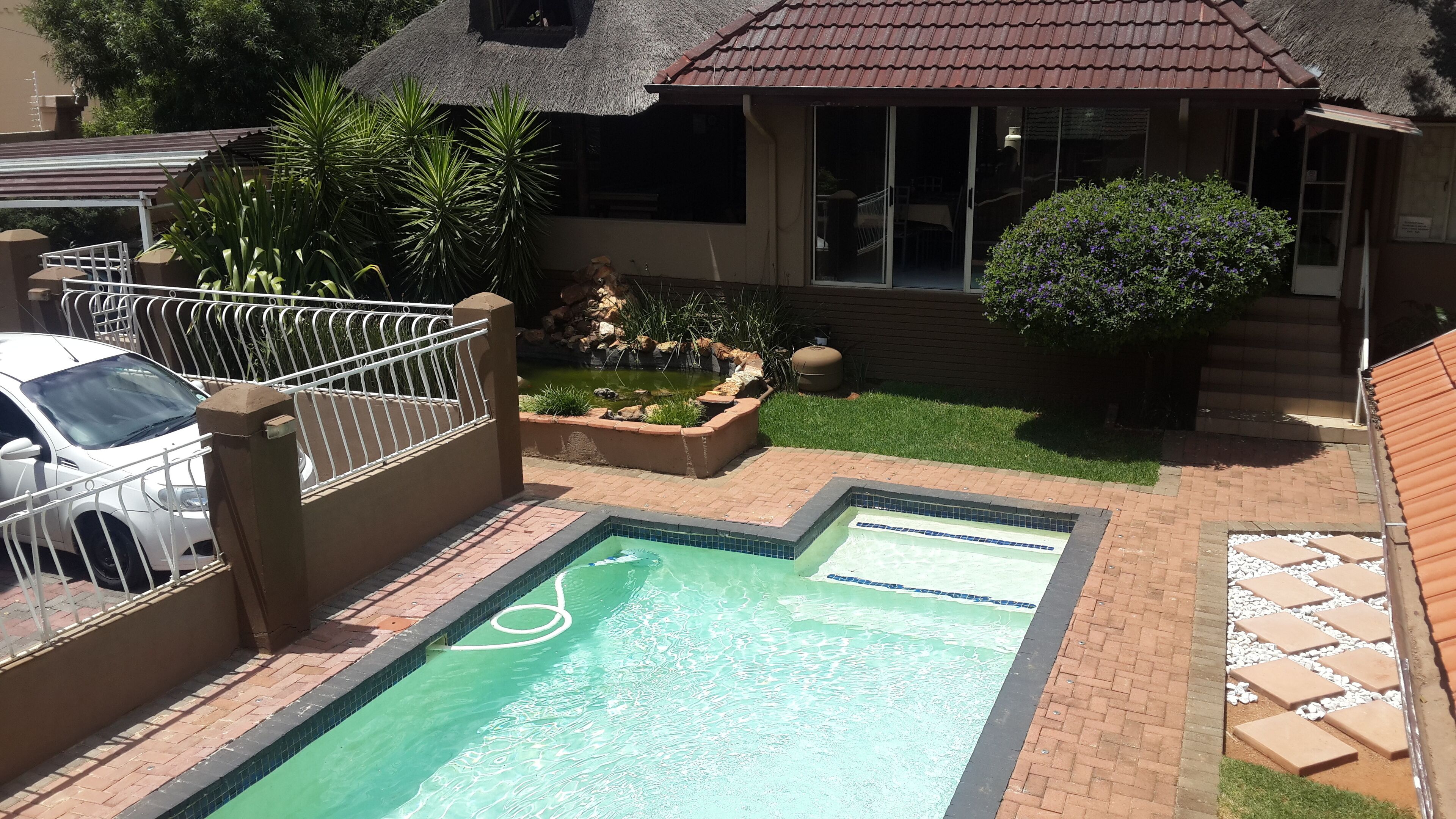 A swimming pool with a tiled border, a fenced area with a car, and a house with a thatched roof.