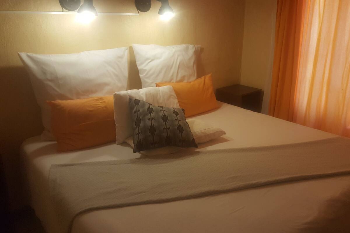 Ein Hotelzimmer mit einem Bett, weißen Kissen, einem orangefarbenen Kissen und einem gemusterten Kissen sowie einem Nachttisch.