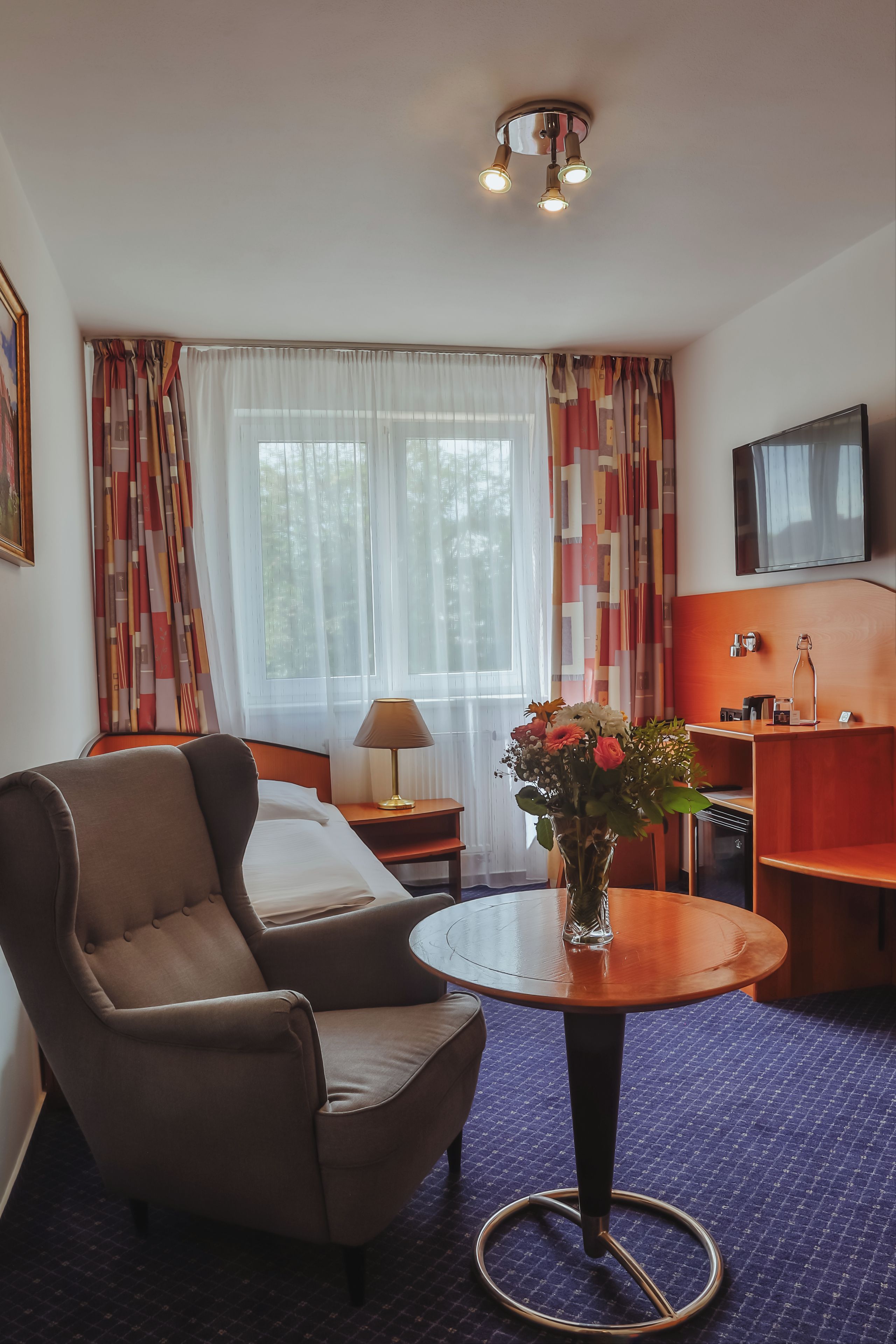 A hotel room with a bed, a chair, a table with a vase of flowers, a desk with a lamp, a television, and a window with curtains.