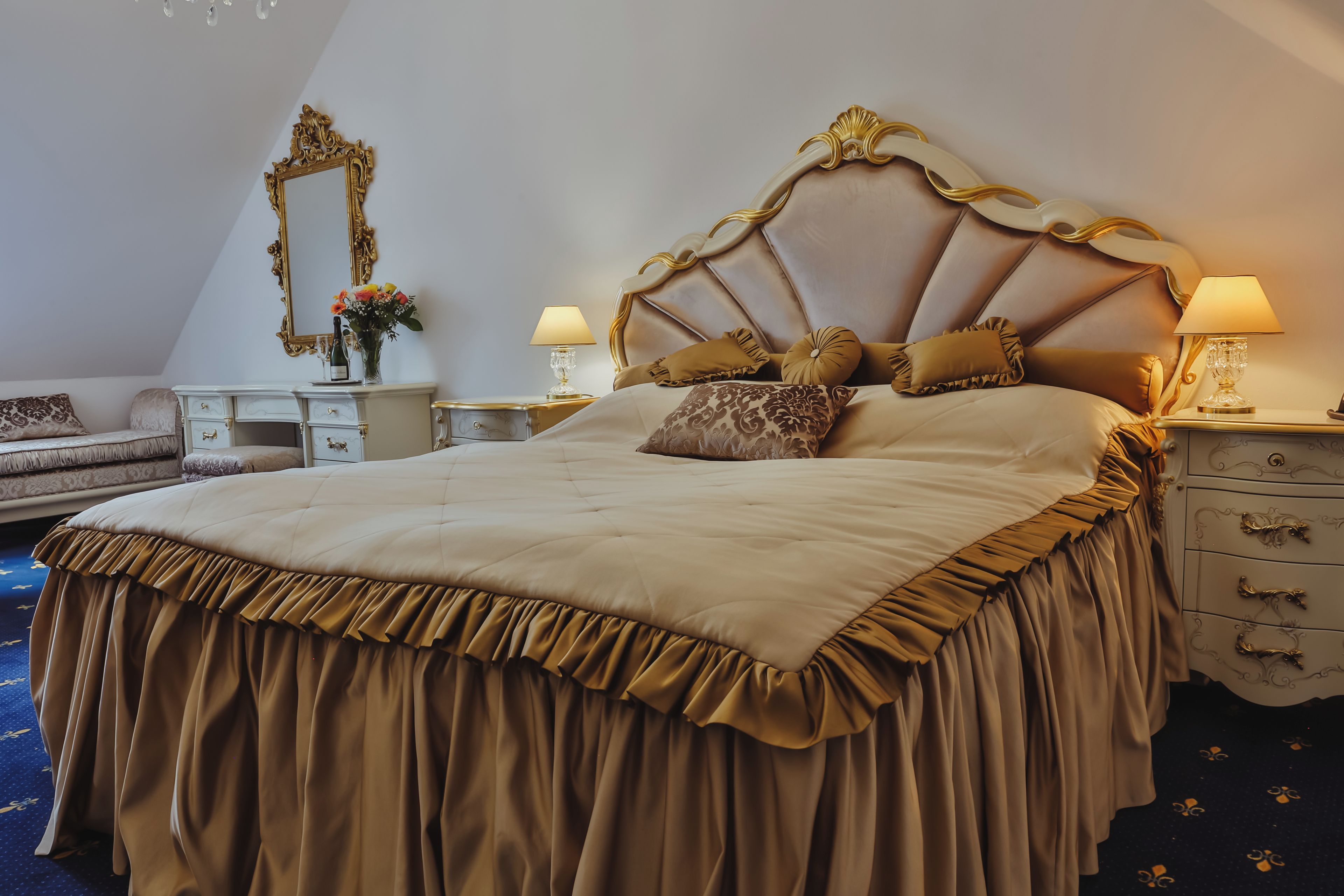A large bed with a decorative headboard, a nightstand with a lamp, a dresser, and a mirror on the wall.