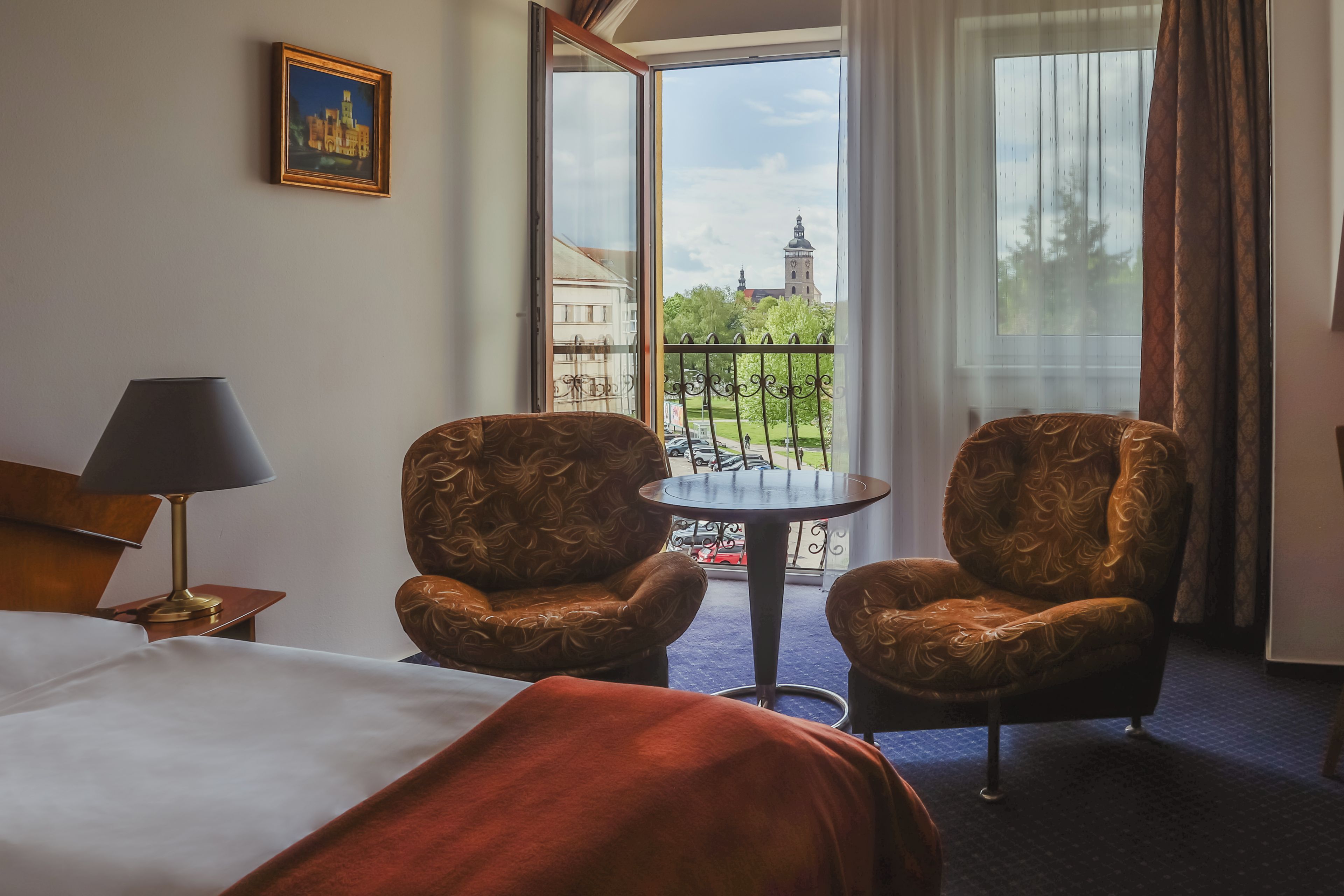 A hotel room with a bed, two armchairs, a small round table, a lamp, and a window with a view of a building and trees.