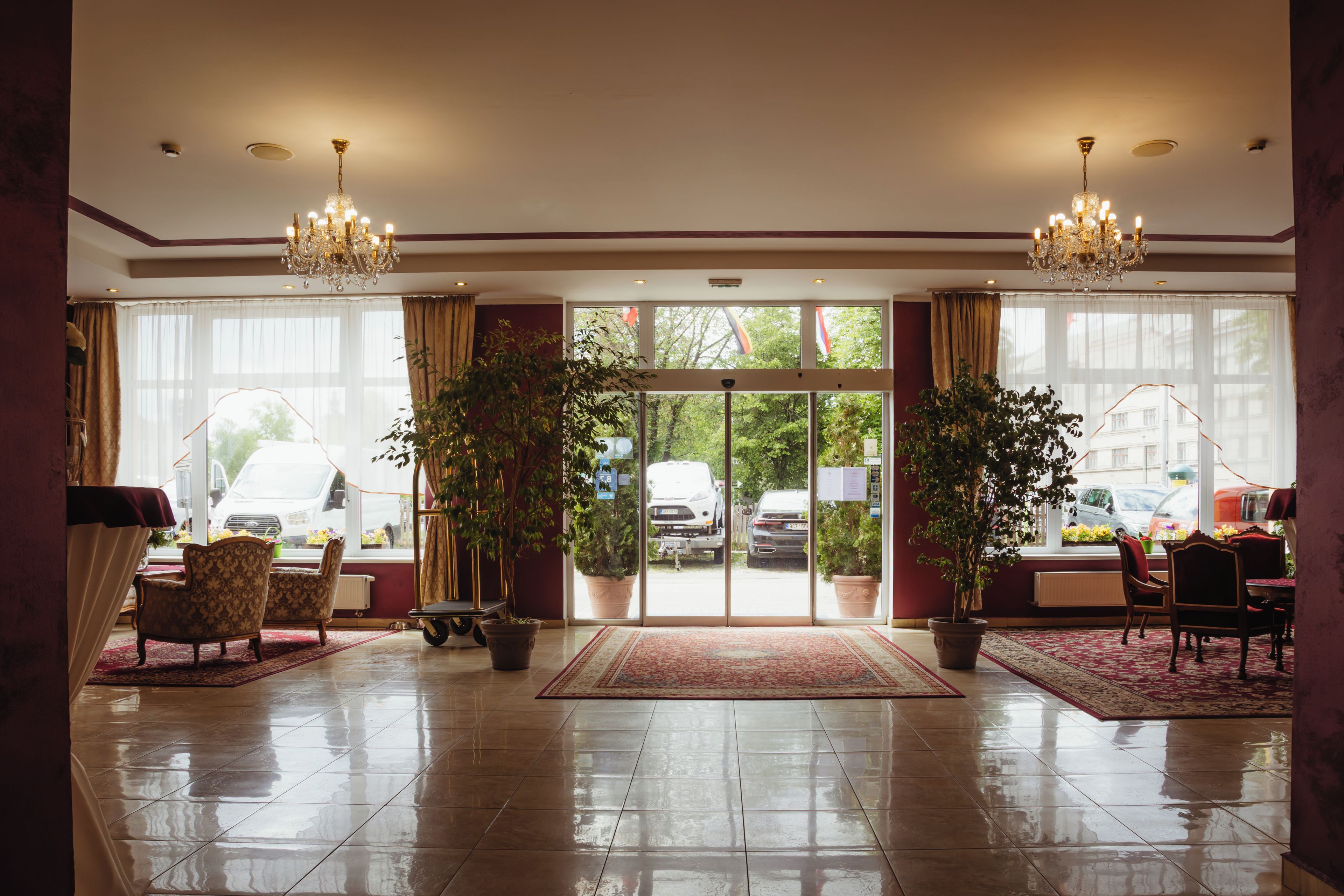 Un hall de hotel con una araña, plantas en macetas, un mostrador de recepción y vistas a la calle a través de puertas de cristal.
