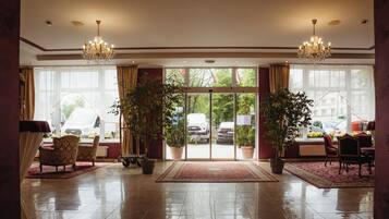 Un hall de hotel con una araña, plantas en macetas, un mostrador de recepción y vistas a la calle a través de puertas de cristal.