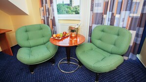 A room with two green upholstered chairs, a small round table with a fruit bowl and a glass of wine, and a view of a building through a window. - Savoy (Ceske Budejovice)