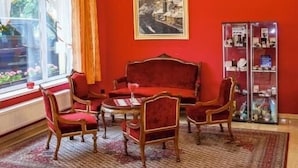 A room with a red wall, a chandelier, a red sofa set, a glass cabinet with items, and a window with curtains. - Savoy (Ceske Budejovice)