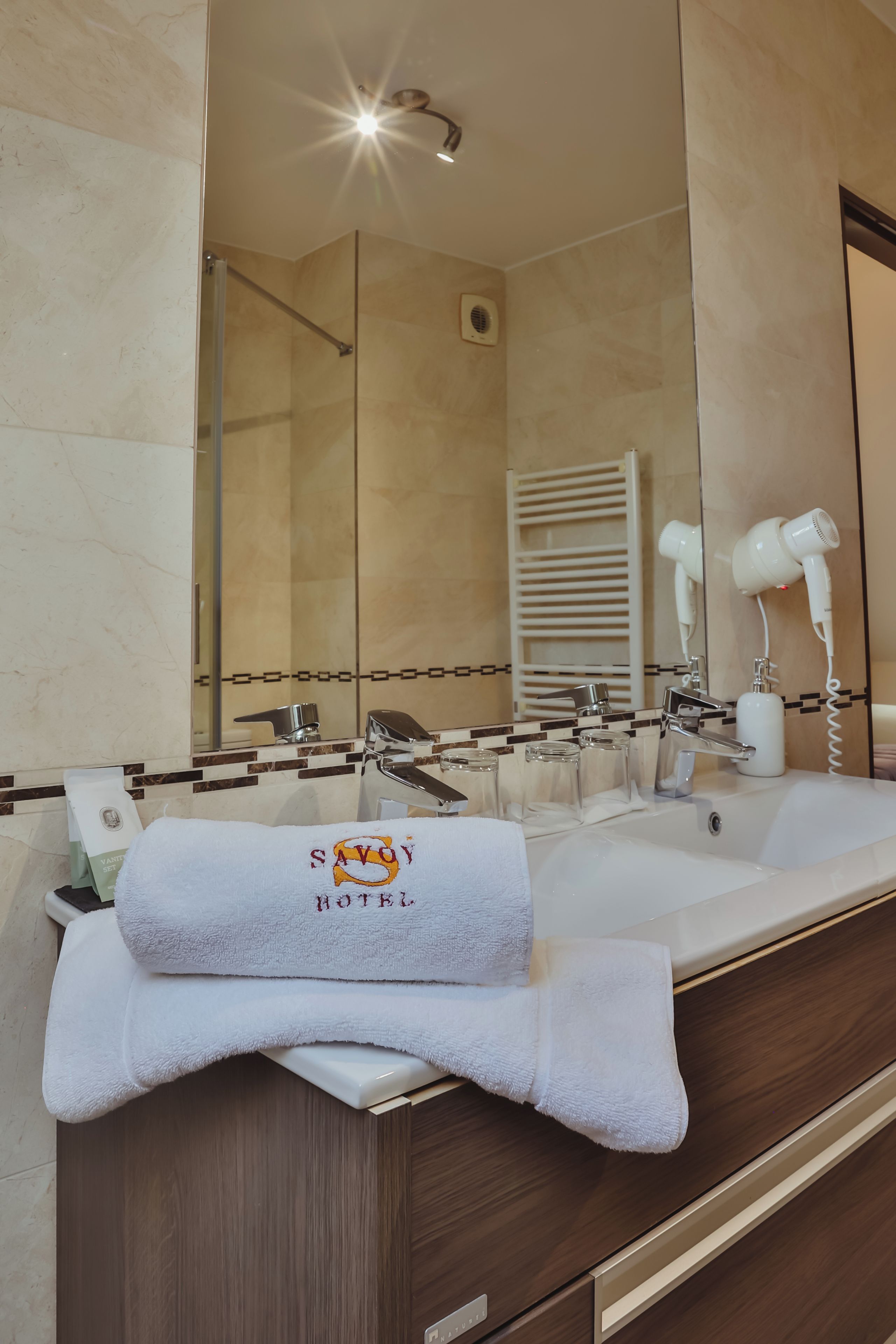Une salle de bain dotée d’un double lavabo, d’une serviette brodée avec le nom « SAVOY HOTEL », d’un sèche-cheveux et d’un miroir qui reflète la pièce.