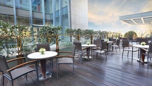 Un patio al aire libre con mesas y sillas redondas, rodeado de follaje y arquitectura moderna.