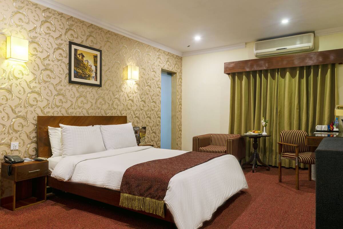A hotel room with a large bed, a desk with a chair, a small table with a lamp, and an air conditioning unit.