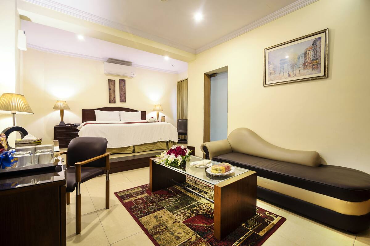 A hotel room with a bed, a sofa, a desk with a lamp, and a painting on the wall.