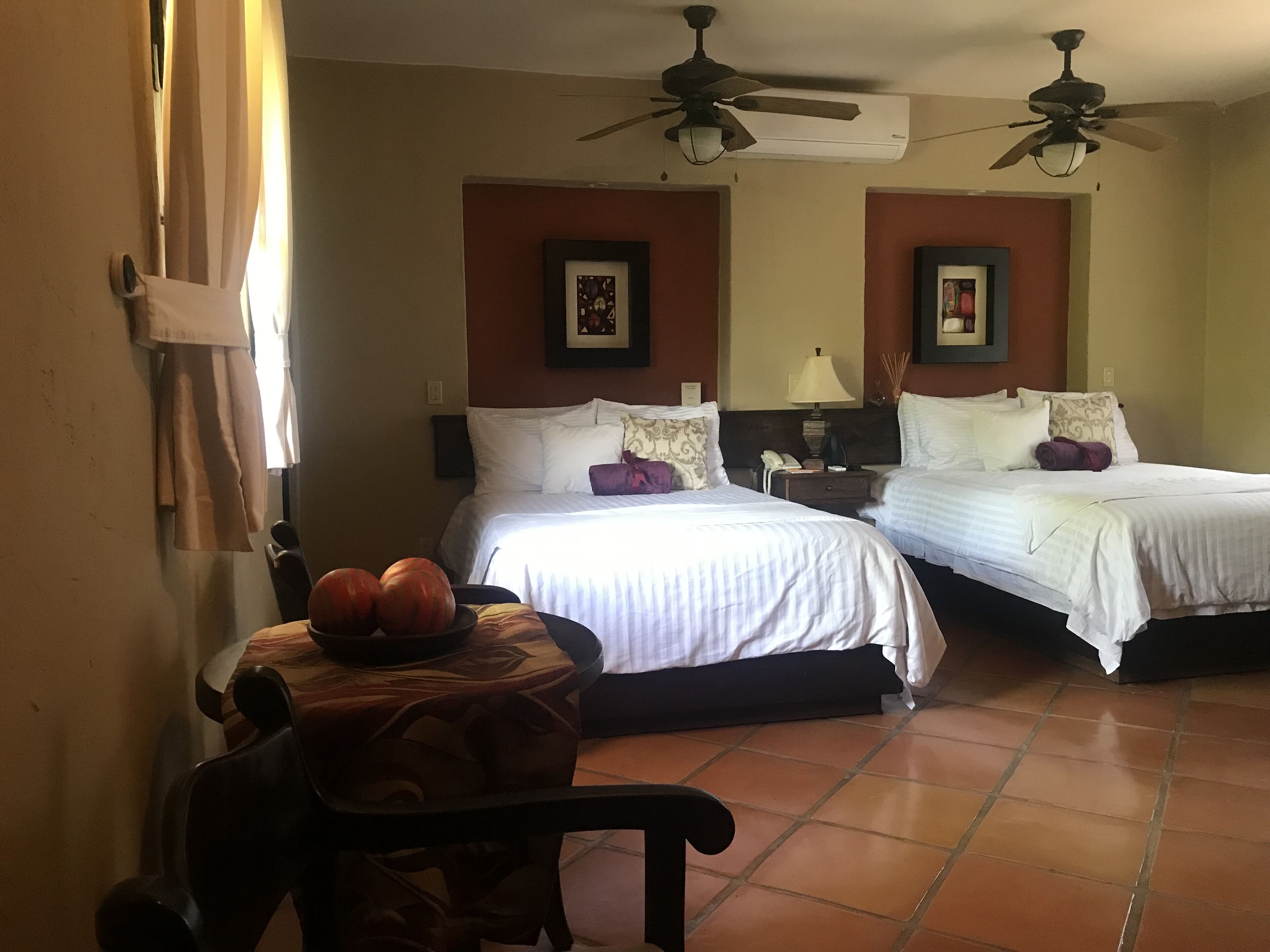 A hotel room with two beds, a chair, a table with fruit, and ceiling fans.
