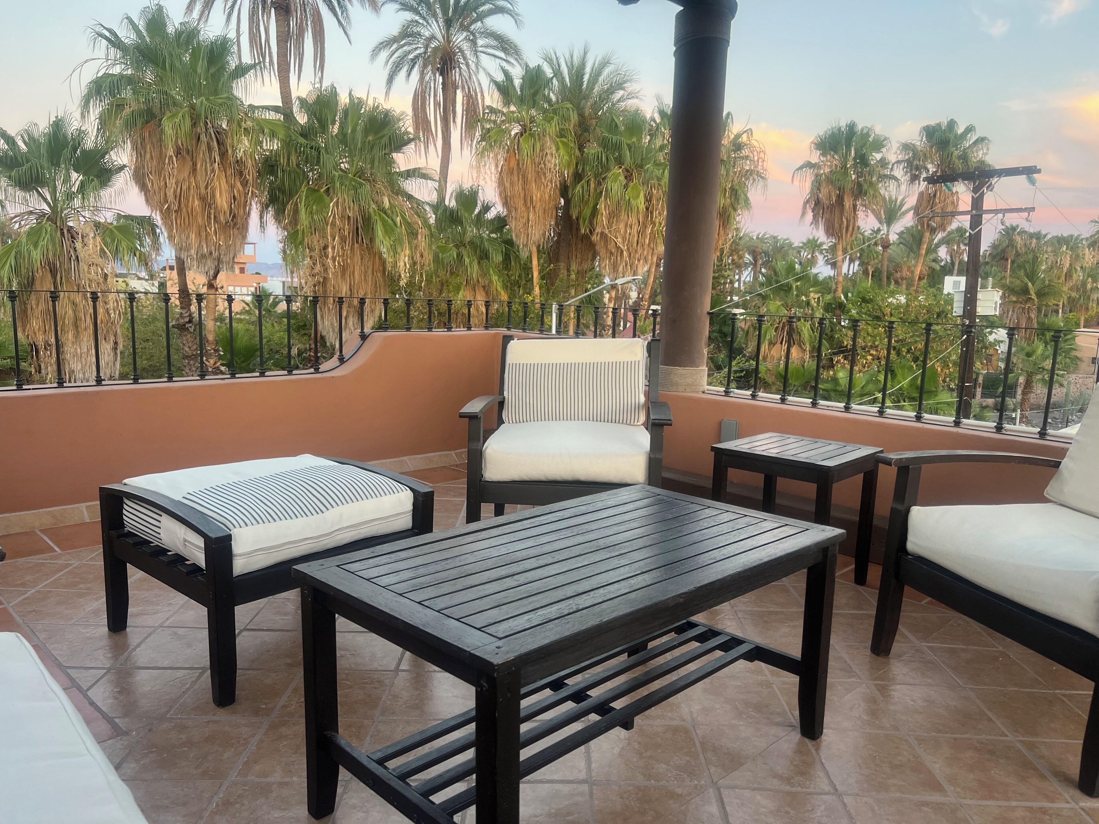 Un balcon agrémenté d’un canapé, de deux fauteuils et d’une petite table, entouré de palmiers et offrant une vue sur le paysage urbain.