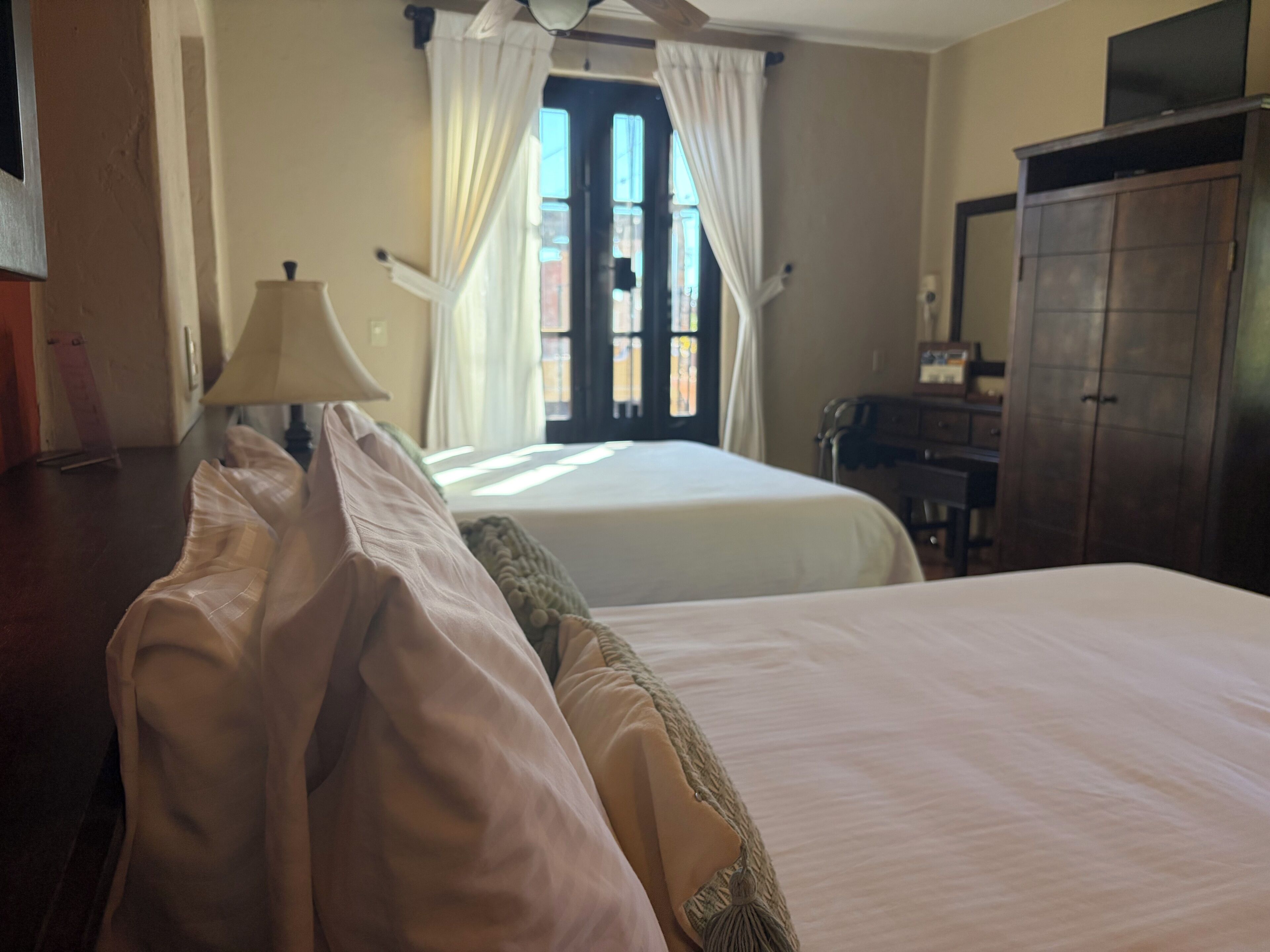 A hotel room with two beds, a wooden wardrobe, a desk, and a window with curtains.