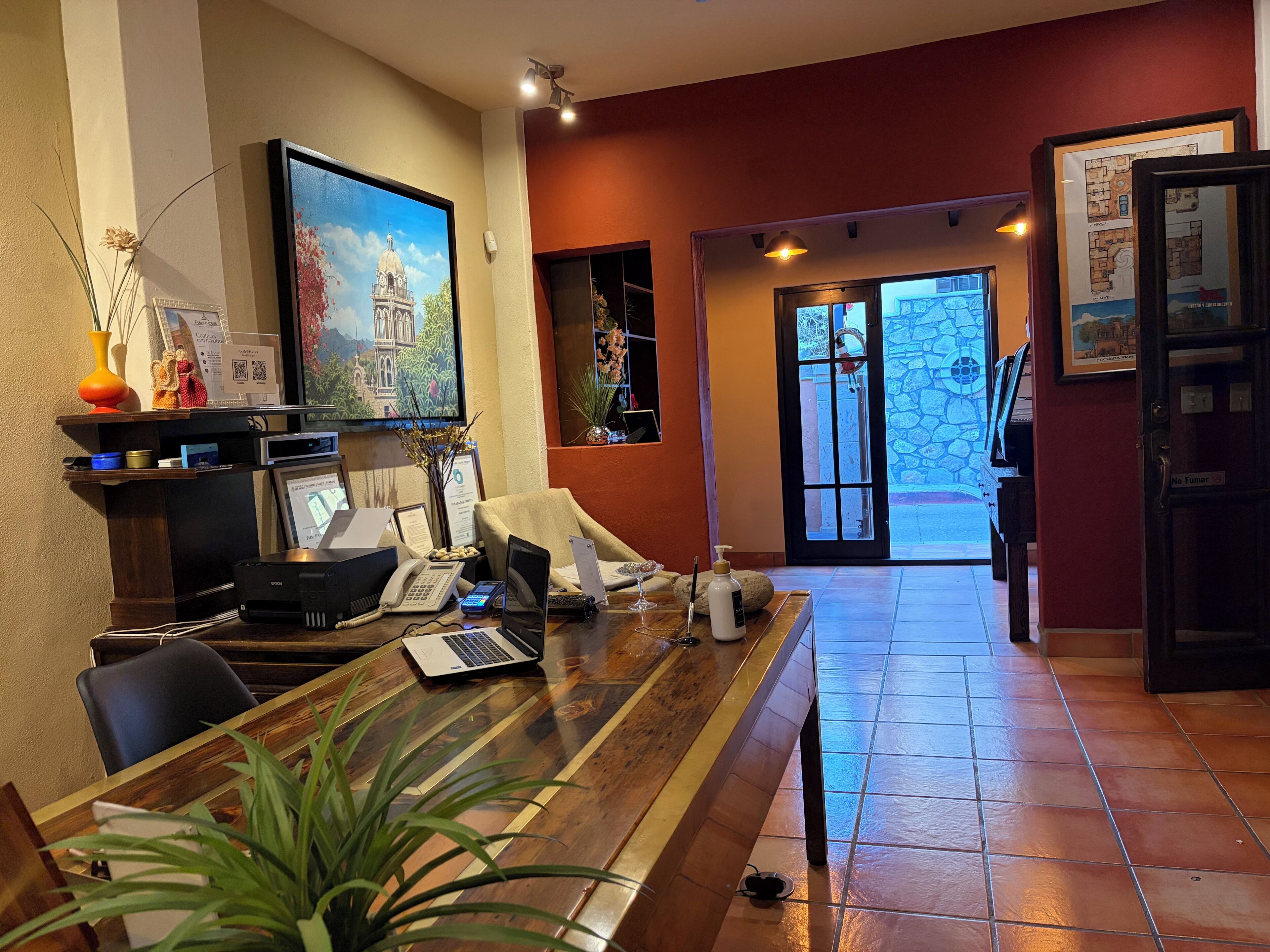 Un mostrador de recepción con una computadora, teléfono y un mapa enmarcado. Hay una planta sobre la mesa y una puerta con un letrero que dice "No Fumar".