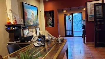 Un mostrador de recepción con una computadora, teléfono y un mapa enmarcado. Hay una planta sobre la mesa y una puerta con un letrero que dice "No Fumar".
