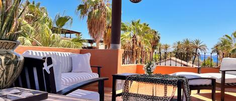 Una terraza en la azotea con mobiliario exterior, un jarrón decorativo y vistas a palmeras y al mar.