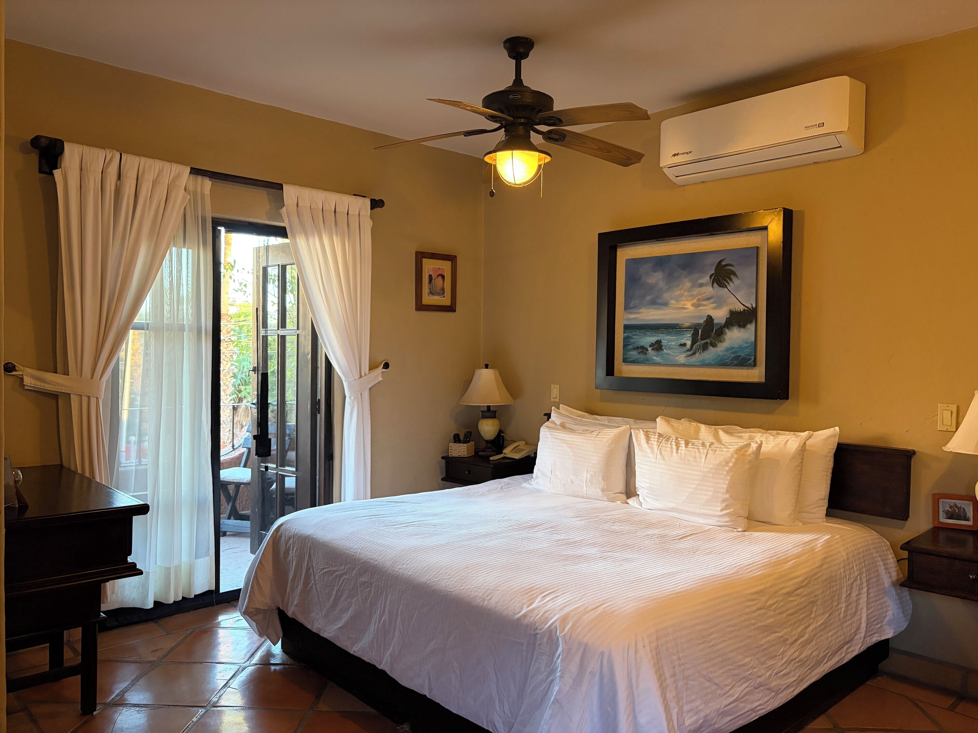 A hotel room with a bed, a ceiling fan, an air conditioner, a painting, a desk, and a chair.