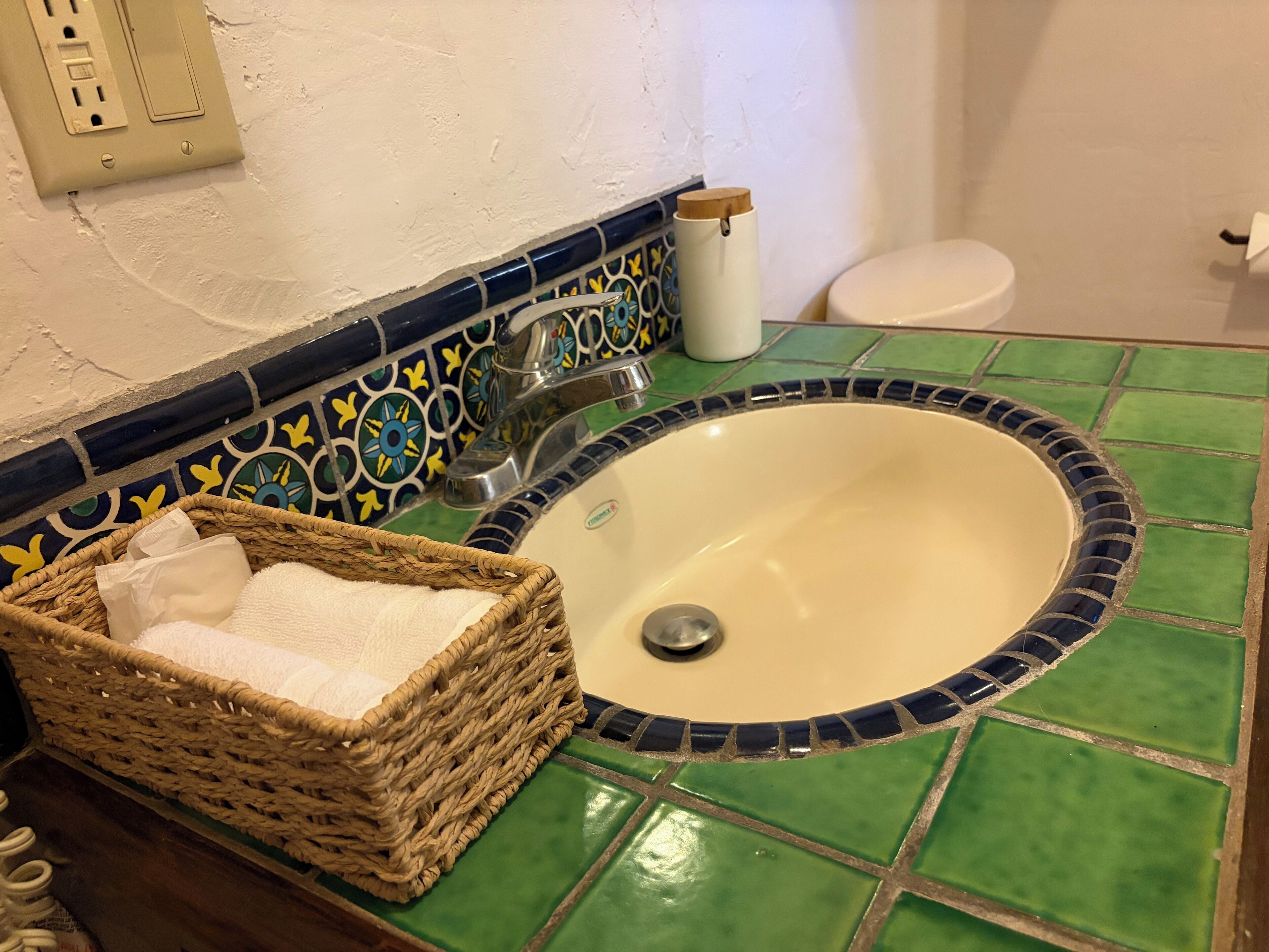 Un baño con un lavamanos de azulejos verdes, un inodoro blanco y una cesta de mimbre con una toalla blanca.