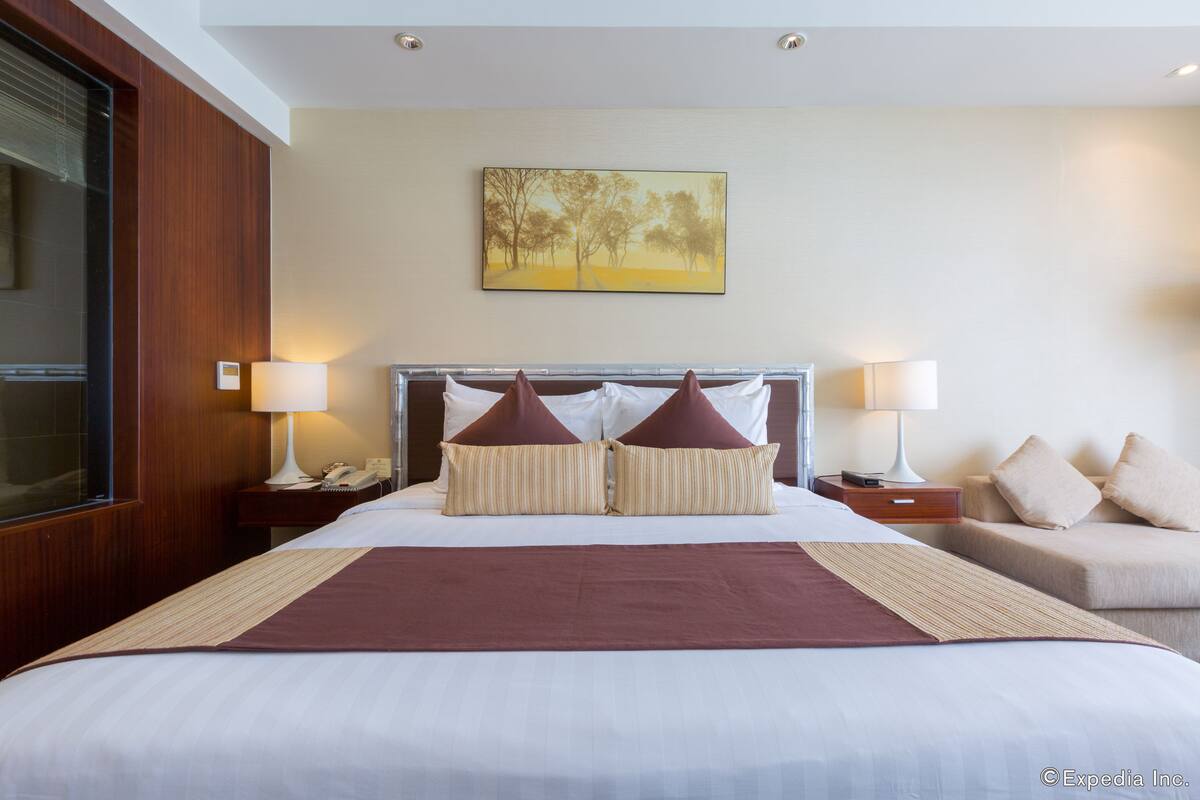 Habitación de hotel con una cama grande, dos mesitas de noche con lámparas, un pouf y un cuadro en la pared.