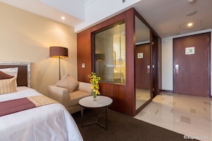 Quarto de hotel com cama, sofá, mesinha com um vaso de flores e uma janela grande com persianas.