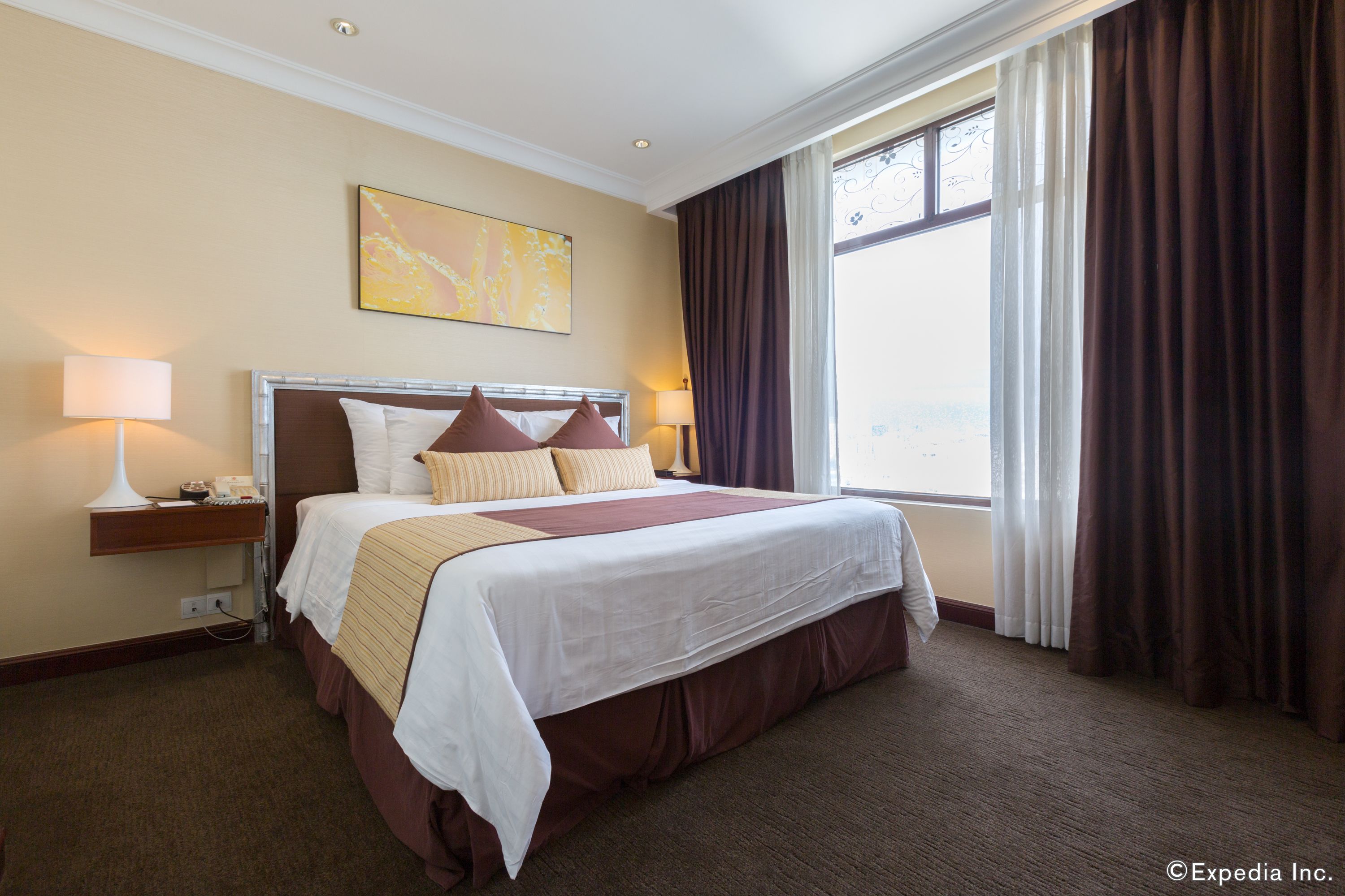 A hotel room with a large bed, a nightstand with a lamp, a window with curtains, and a painting on the wall.