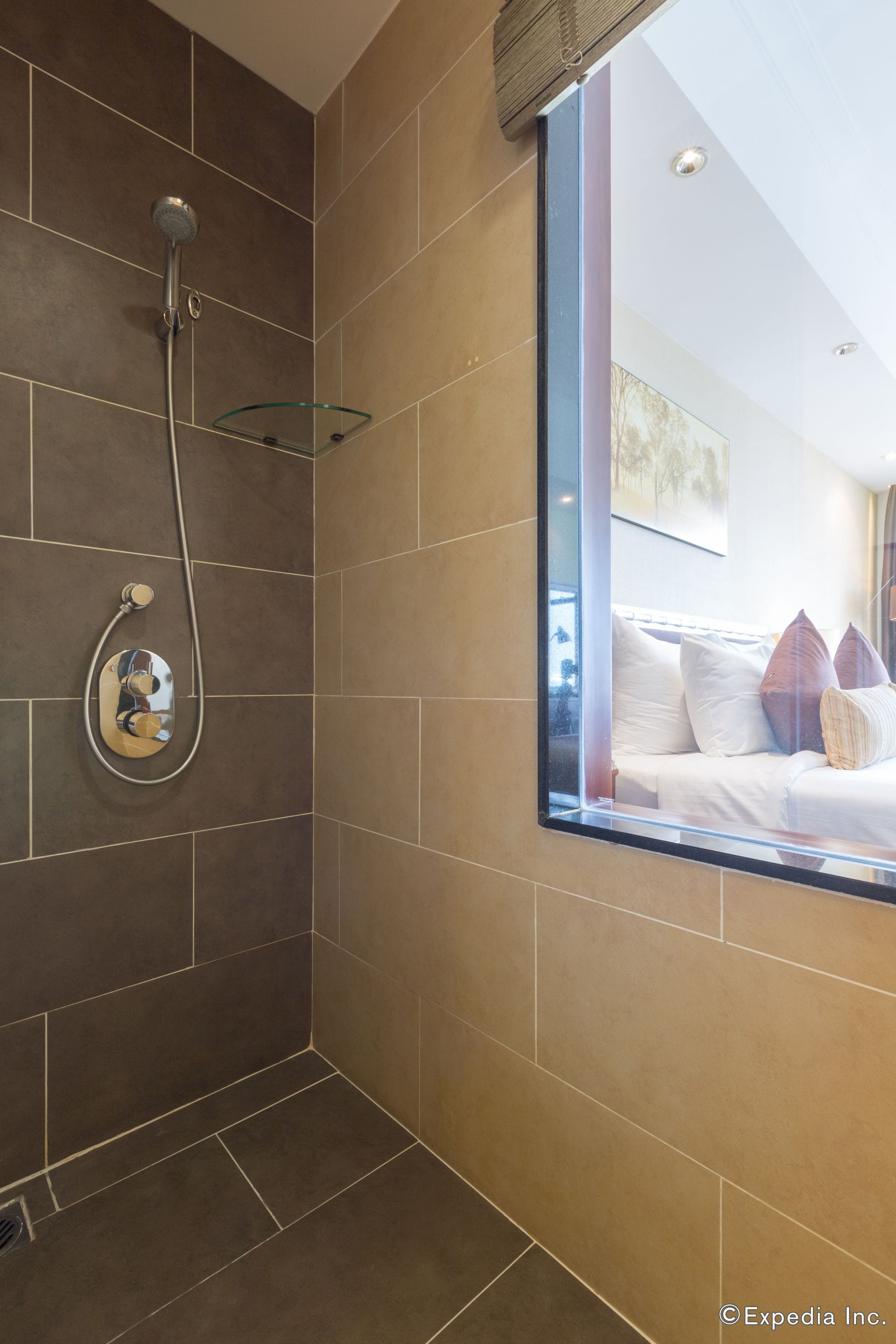 A modern bathroom with a shower, a glass-enclosed window, and a bed visible through the window.