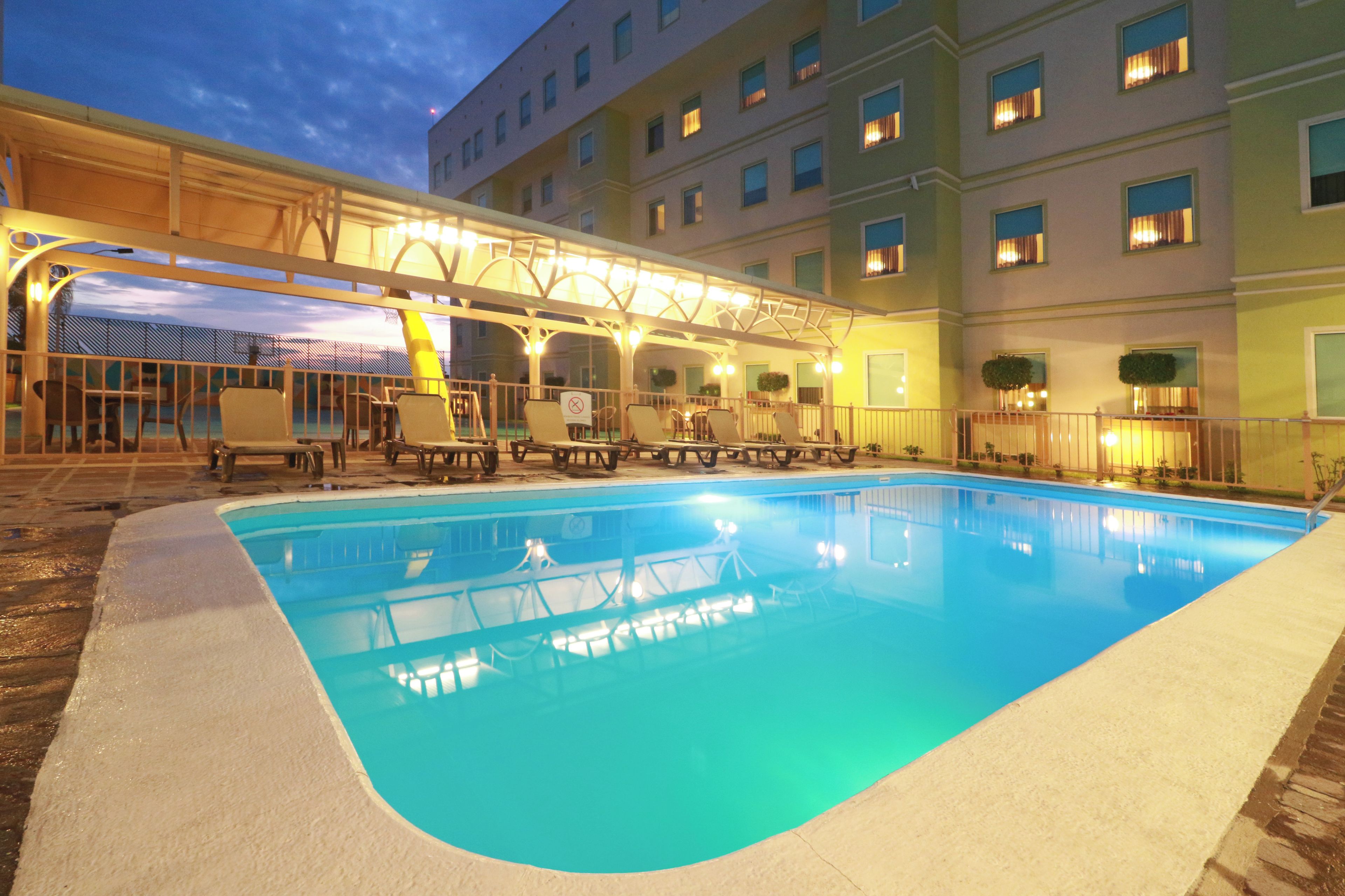 A swimming pool with lounge chairs and a covered seating area, surrounded by a building with multiple windows.