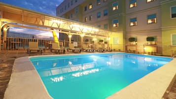 A swimming pool with lounge chairs and a covered seating area, surrounded by a building with multiple windows.