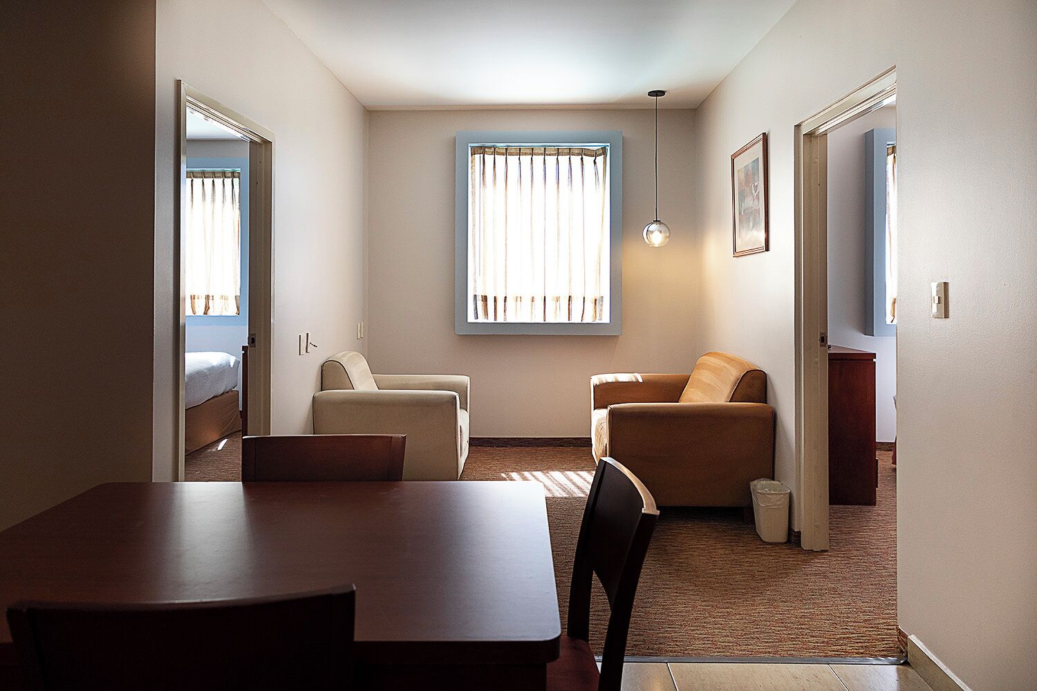 A compact hotel room with a dining table, two armchairs, a sofa, a bed, and a large window with curtains.