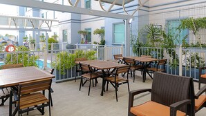 Eine Dachterrasse mit Holztischen und -stühlen, umgeben von Pflanzen und Blick auf einen Pool.