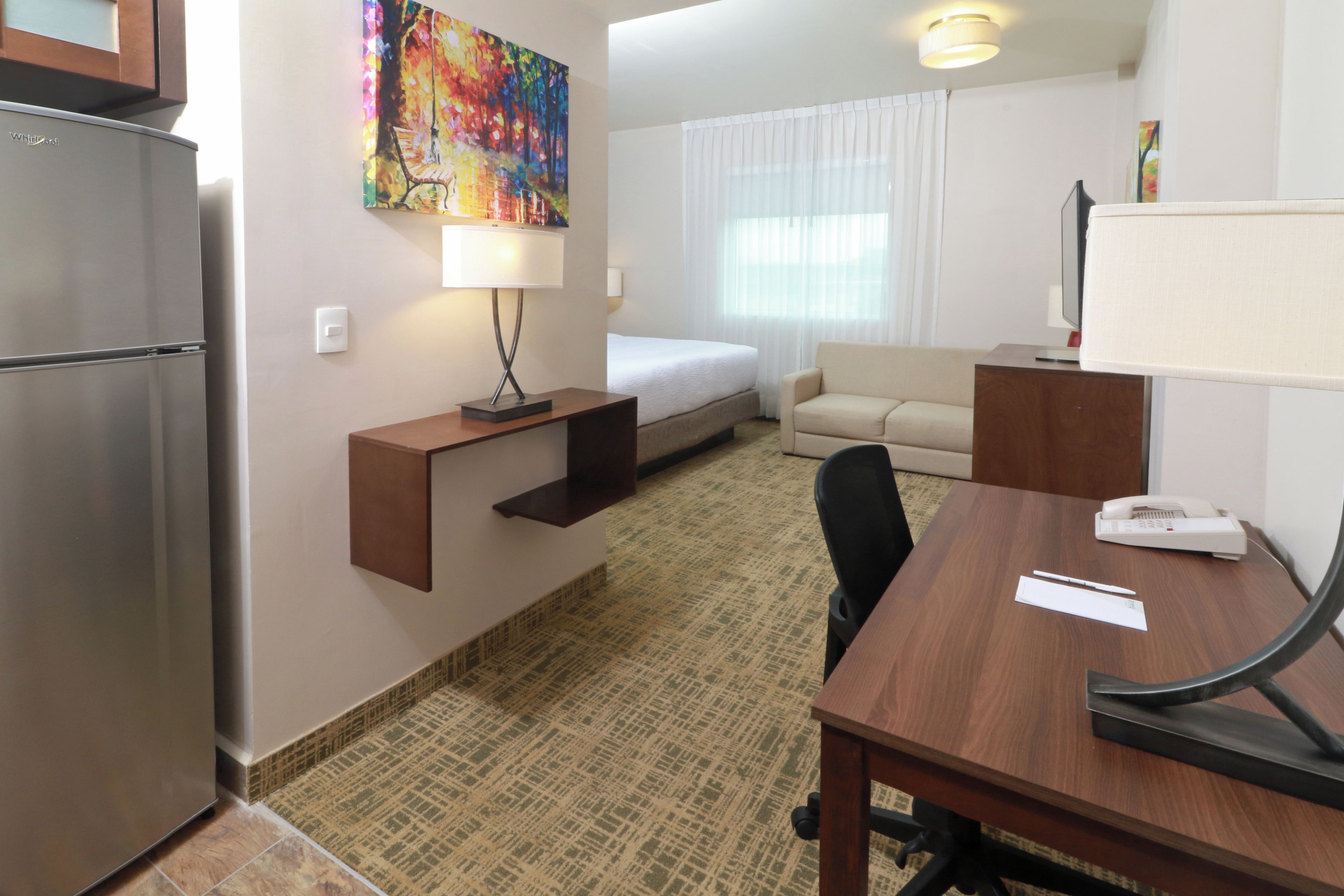 Habitación de hotel con cama, escritorio, silla, refrigerador, lámpara y un cuadro en la pared.