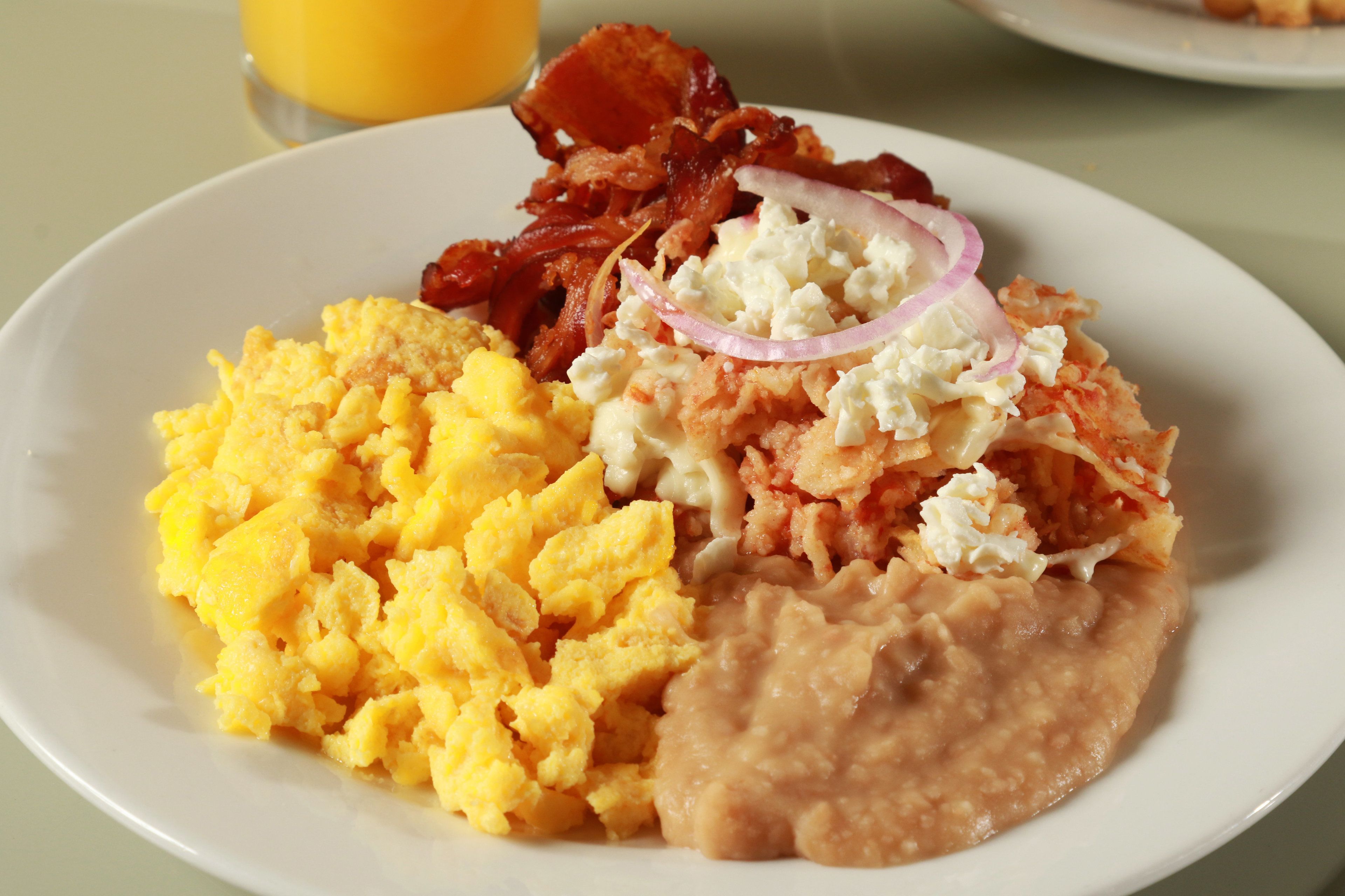 A plate of scrambled eggs, bacon, beans, and a slice of red onion.