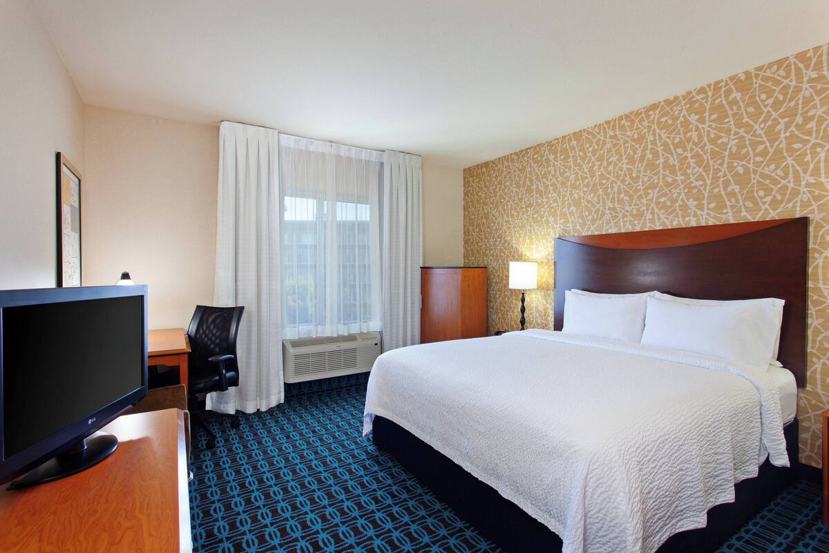Une chambre d’hôtel avec un lit, un bureau avec un ordinateur, une chaise, une télévision et une fenêtre avec des rideaux.