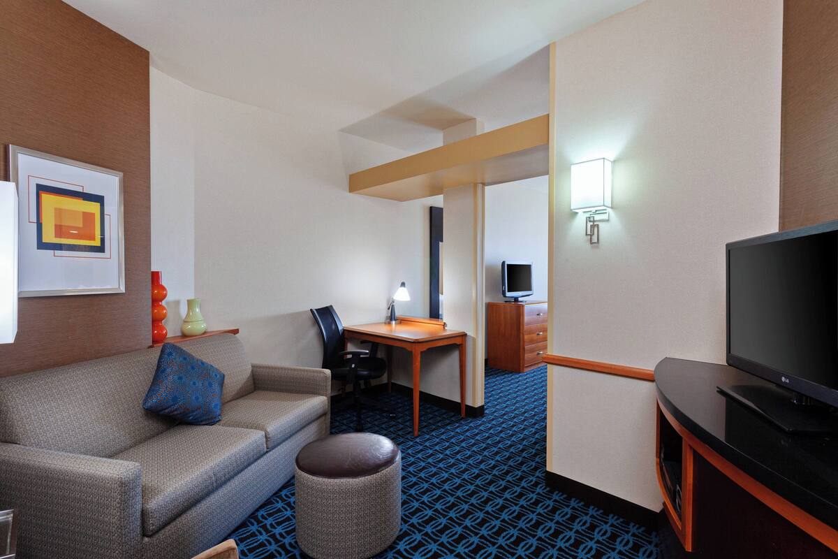 Une chambre d’hôtel équipée d’un canapé, d’un pouf, d’un bureau, d’une chaise, d’une télévision et d’une lampe.