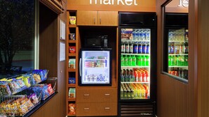 Un pequeño espacio comercial con un refrigerador lleno de bebidas y aperitivos, un microondas y estantes con diversos productos envasados.