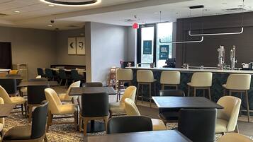 A modern hotel lobby with a mix of dining tables and chairs, a reception desk, and a large window providing natural light.