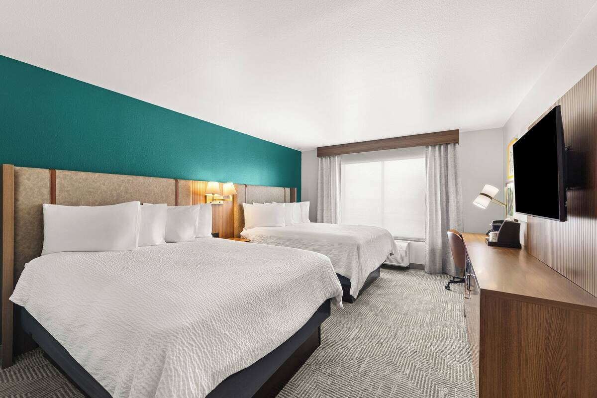 Ein Hotelzimmer mit zwei Betten, einem Schreibtisch, einem Fernseher und einem Fenster mit Vorhängen.