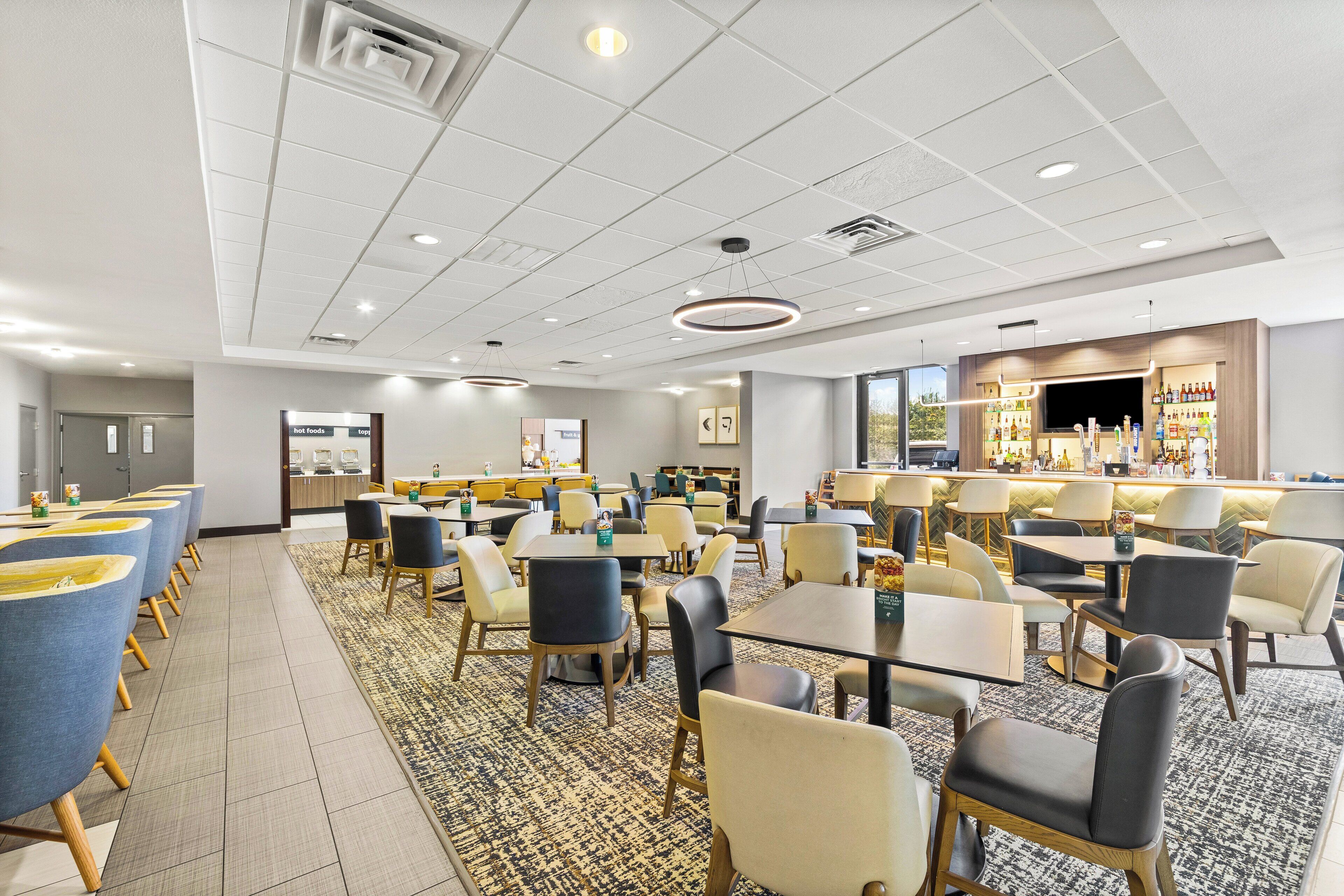 A hotel dining area with multiple tables and chairs, a bar area with a TV, and a reception desk.