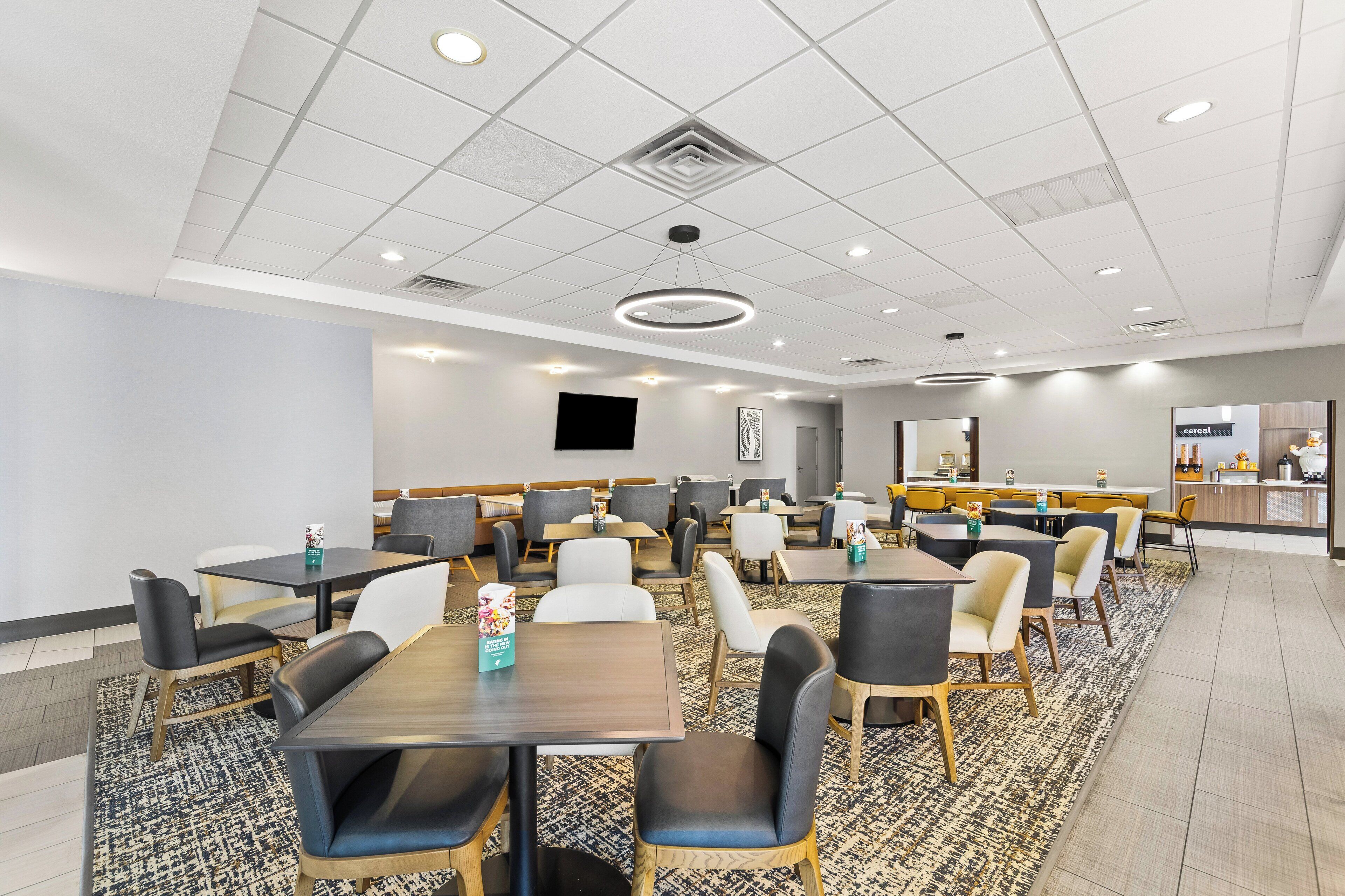 A modern hotel dining area with multiple tables and chairs, a patterned rug, and a counter with a menu.