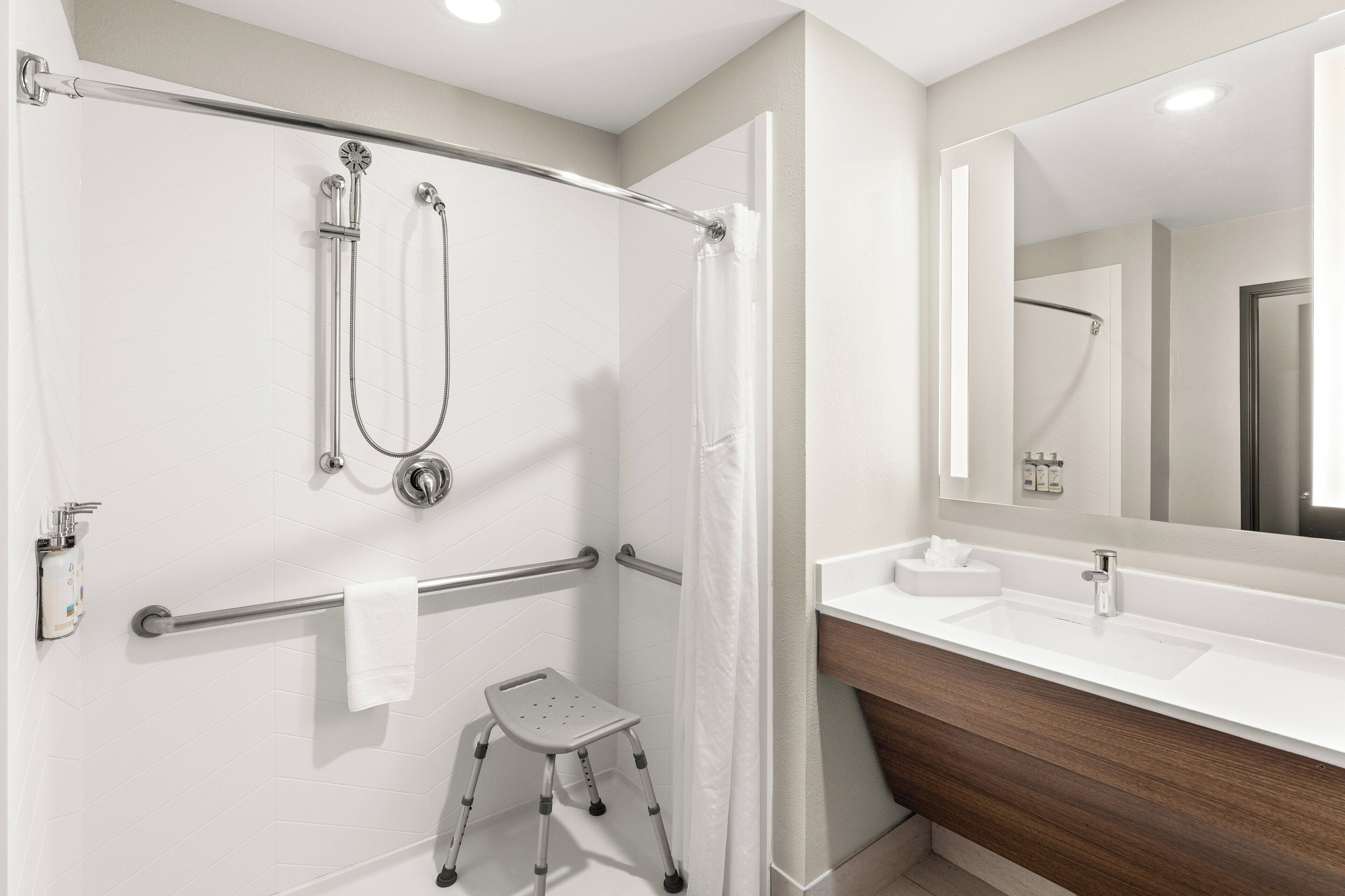 Un baño con ducha, barra de apoyo y lavabo con espejo.