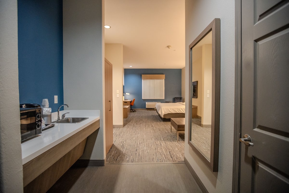 A hotel room with a kitchenette, a bed, a desk, and a bathroom.