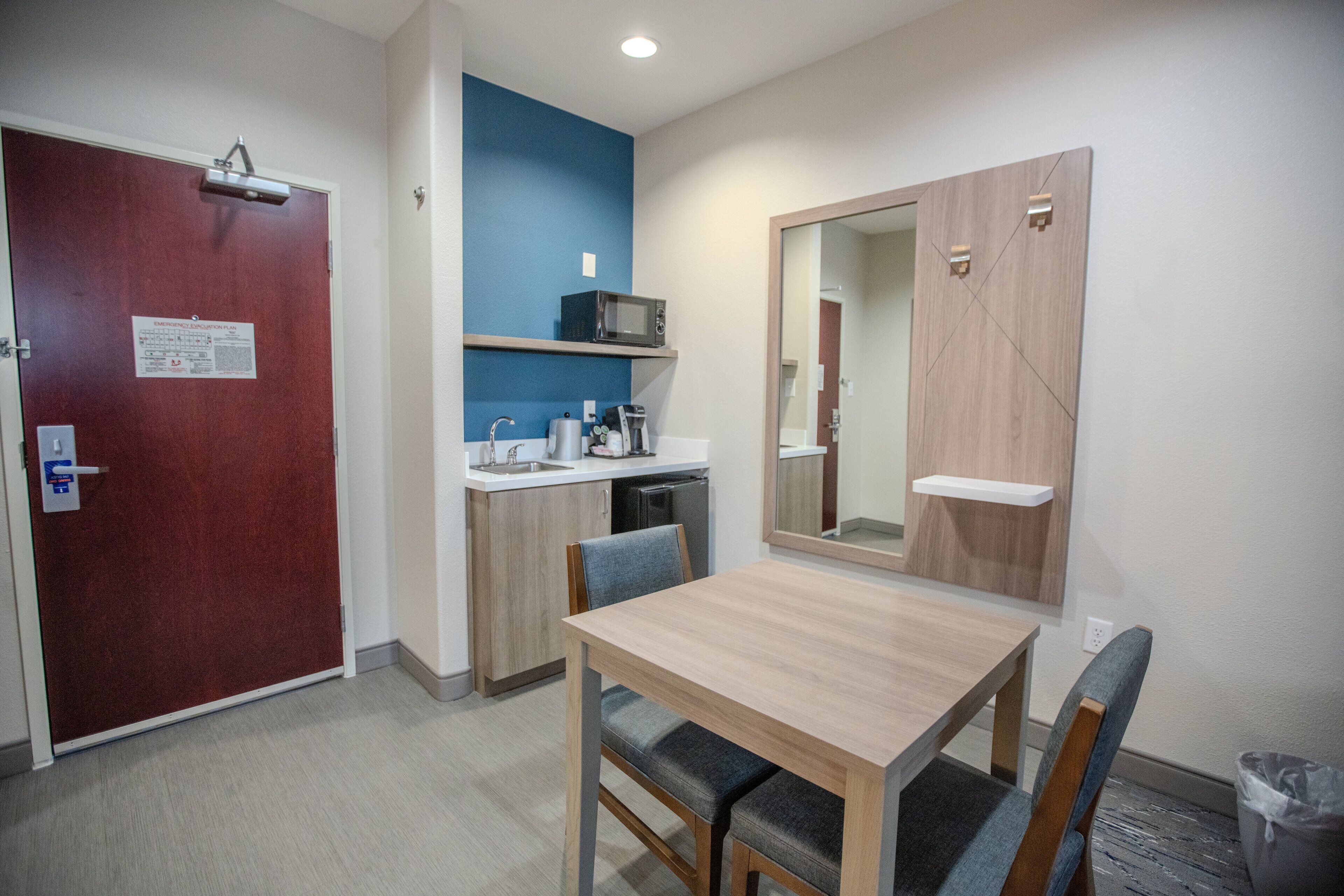 A compact hotel room with a wooden dining table, a microwave, a sink, and a mirror.