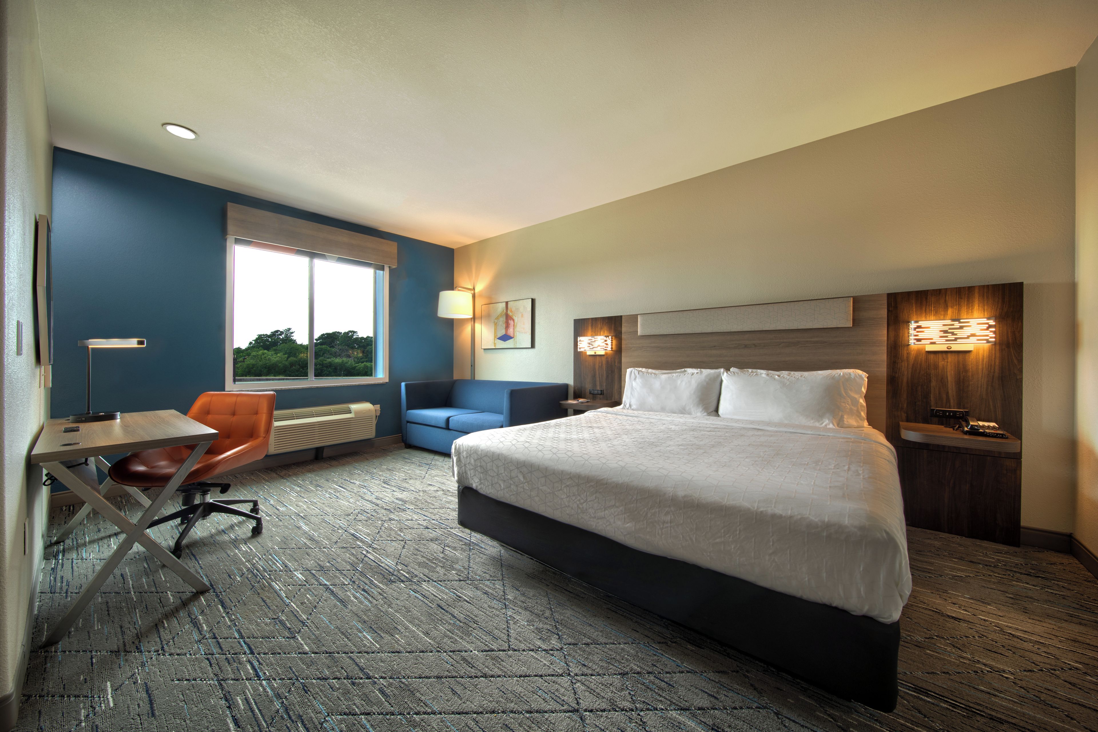 A hotel room with a bed, a desk with a chair, a sofa, and a window with a view of trees.