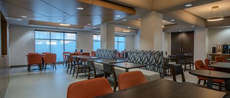 A spacious indoor area with multiple tables and chairs, large windows, and a modern ceiling design.