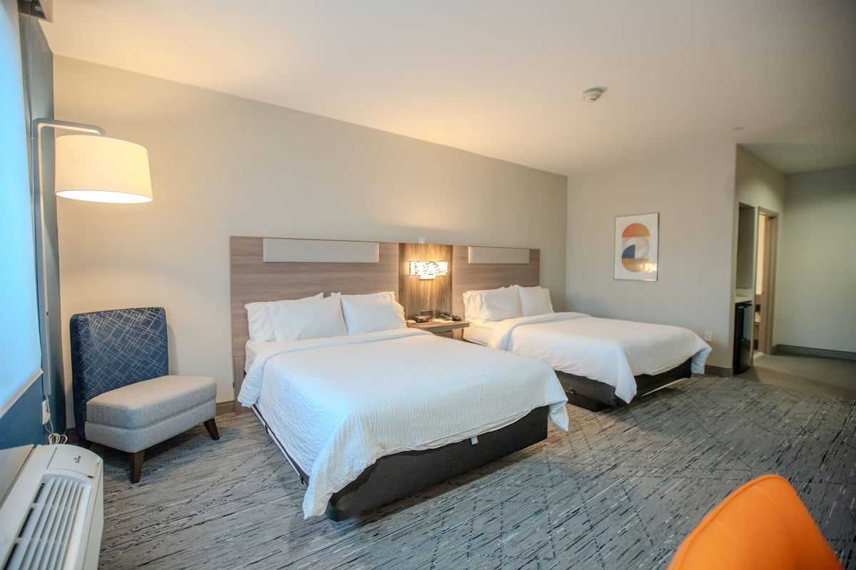 A hotel room with two beds, a chair, a desk, and a lamp.