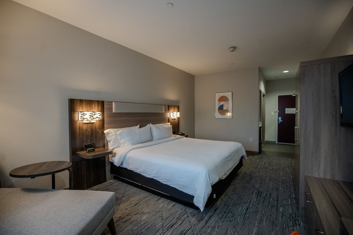 A hotel room with a bed, bedside tables, a chair, a TV, and a door leading to another room.