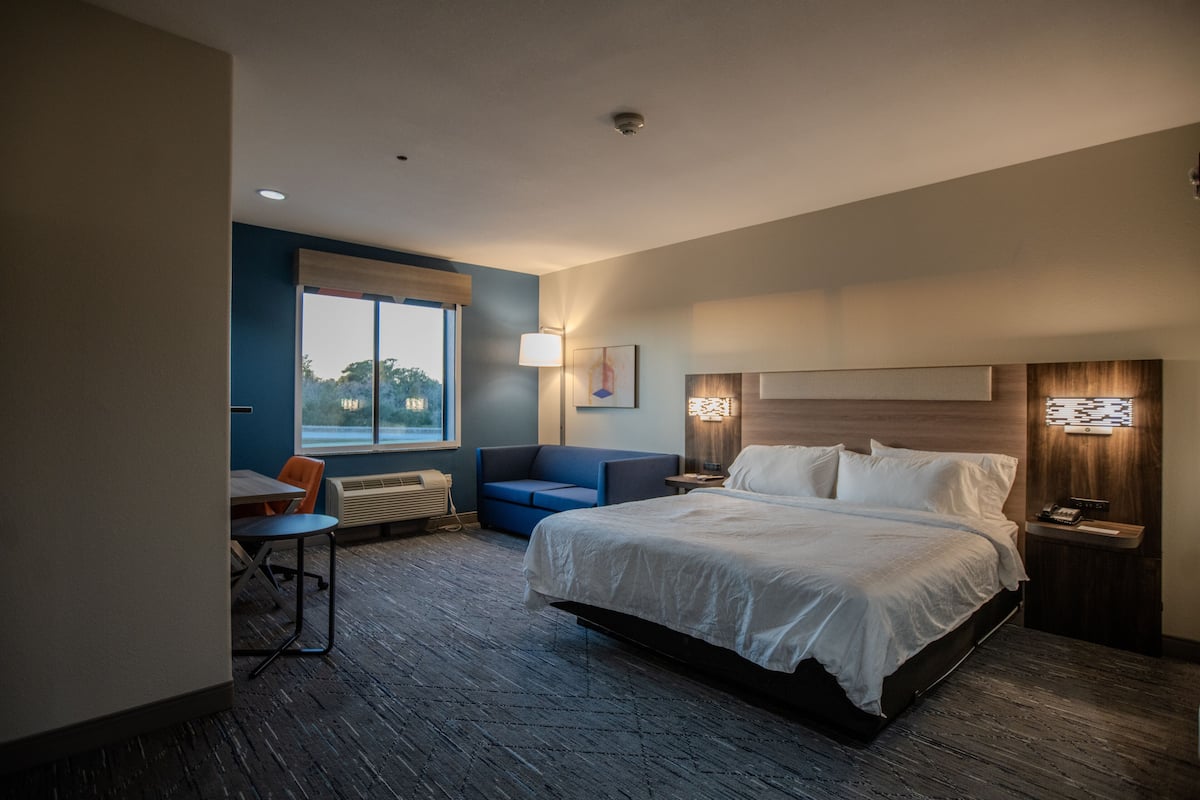 A hotel room with a large bed, a desk with a chair, a sofa, and a window with a view of trees.