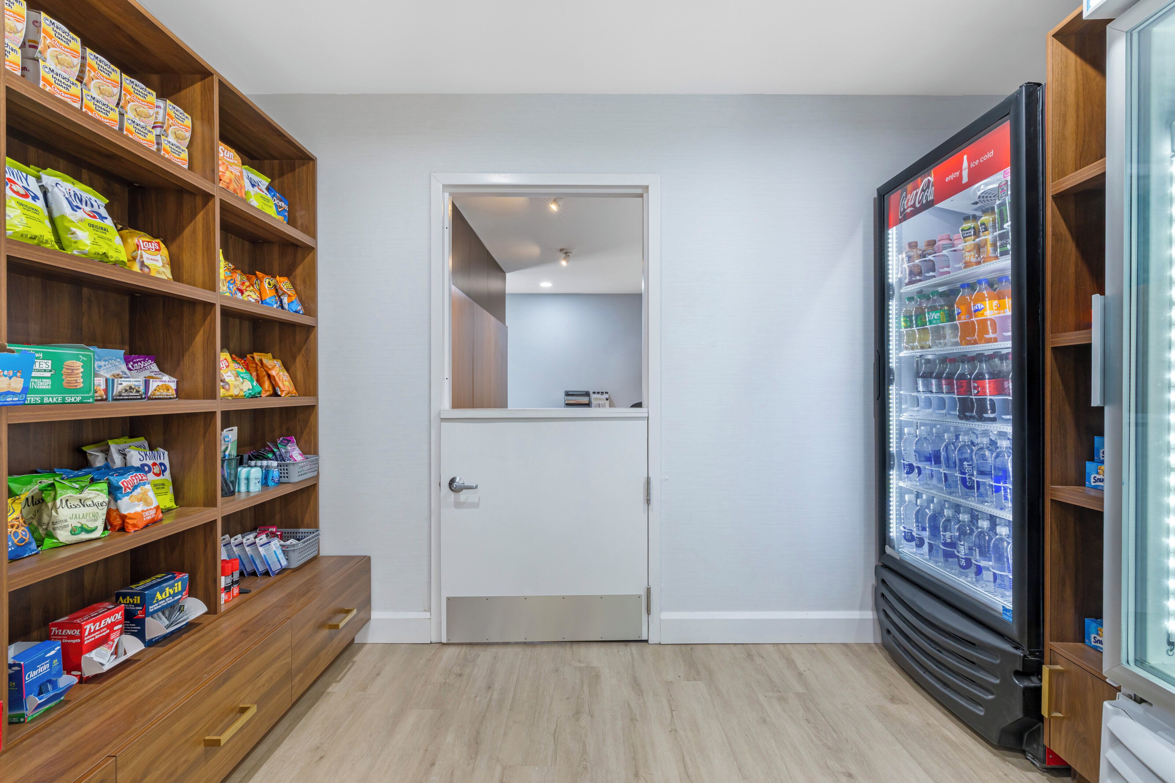 A storage room with shelves stocked with snacks and beverages, a door leading to another room, and a refrigerator with drinks.