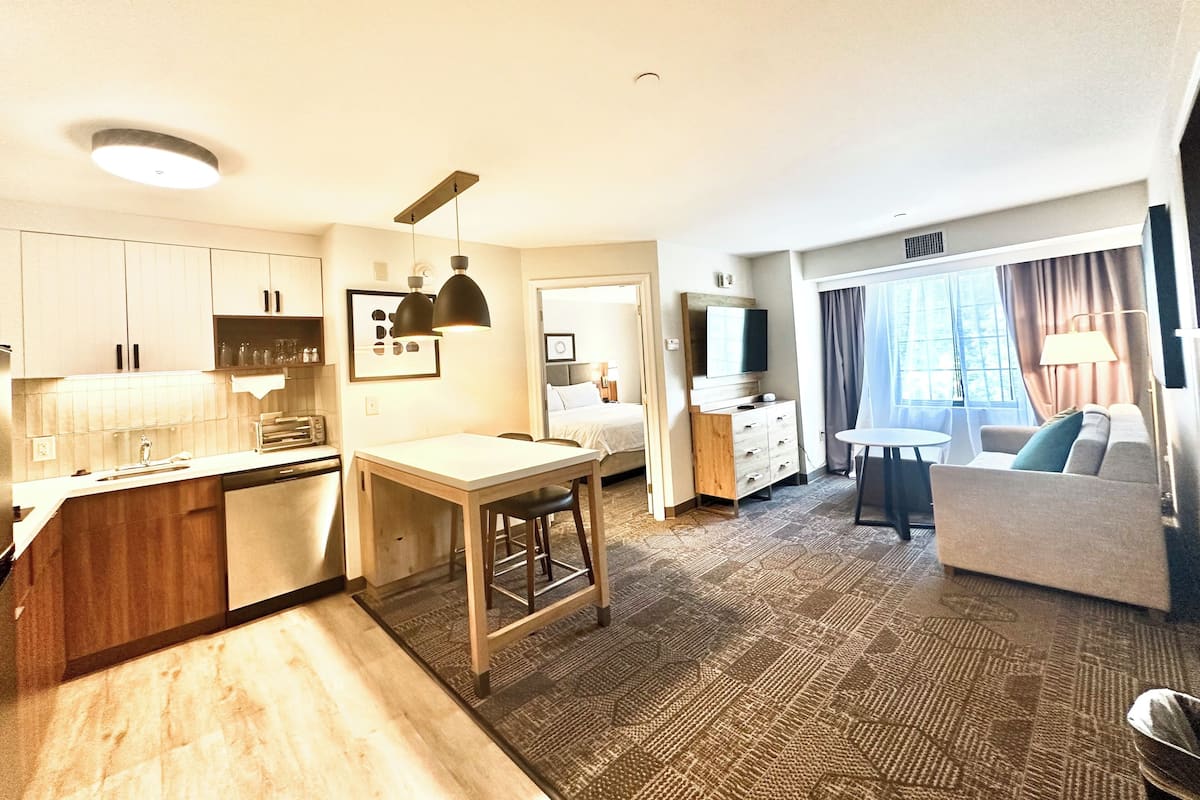 A compact hotel room with a kitchenette, a dining area, a sofa, and a TV.