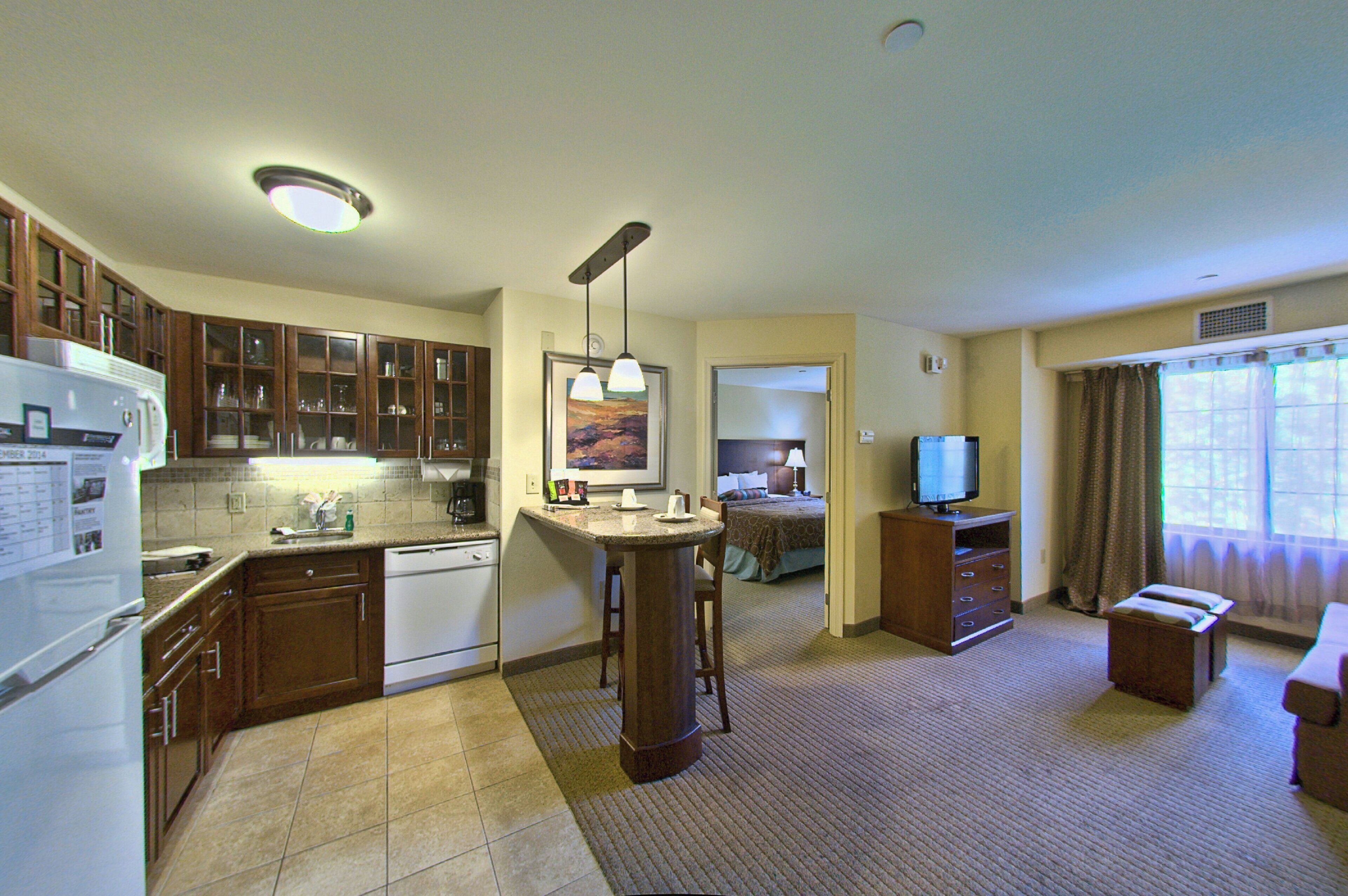 A compact hotel room with a kitchenette, a dining area, a television, and a window with curtains.